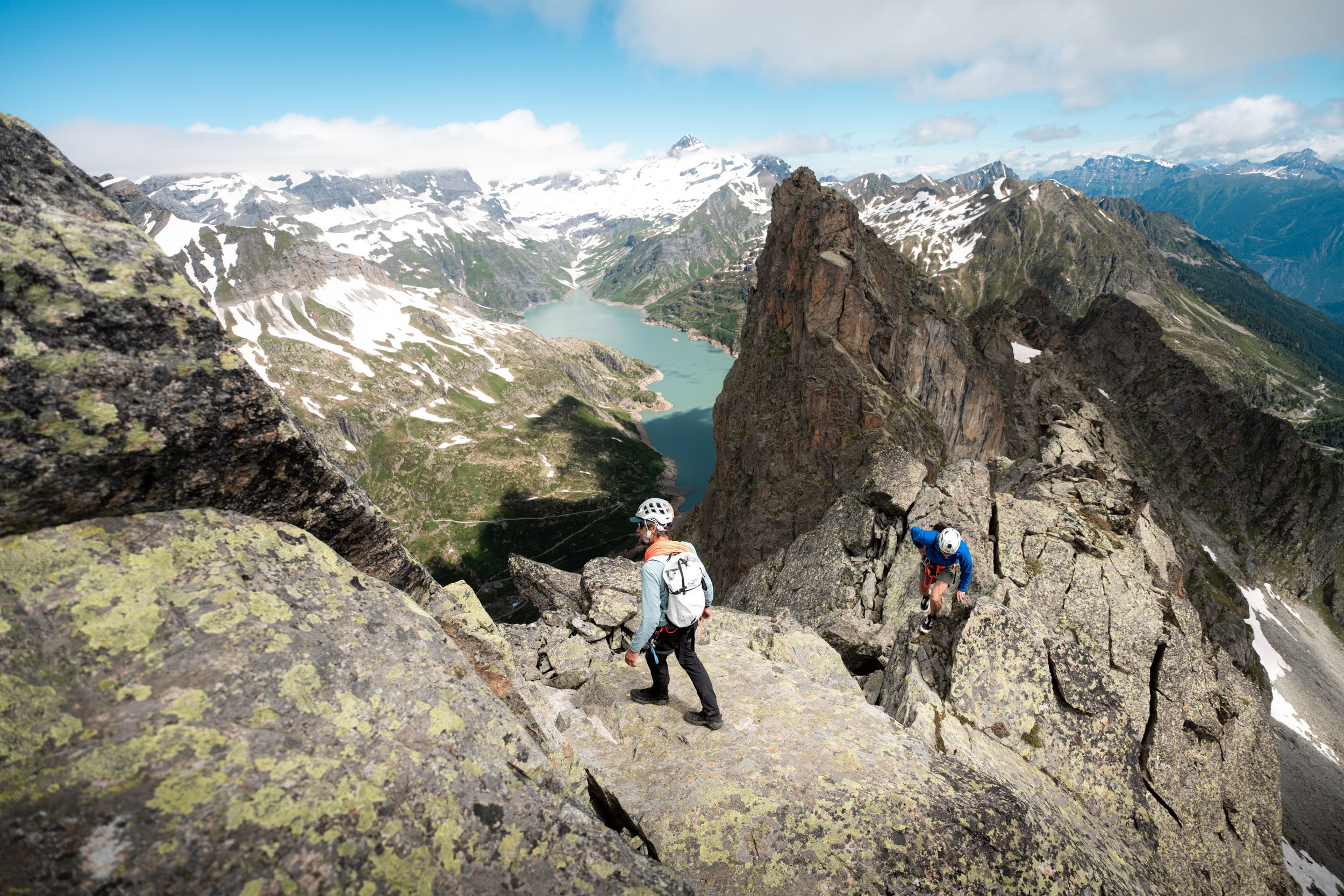 Two climbers with helmets and backpacks ascending a rocky mountain with a scenic view of a lake, snow-capped peaks, and a cloudy sky in the background.