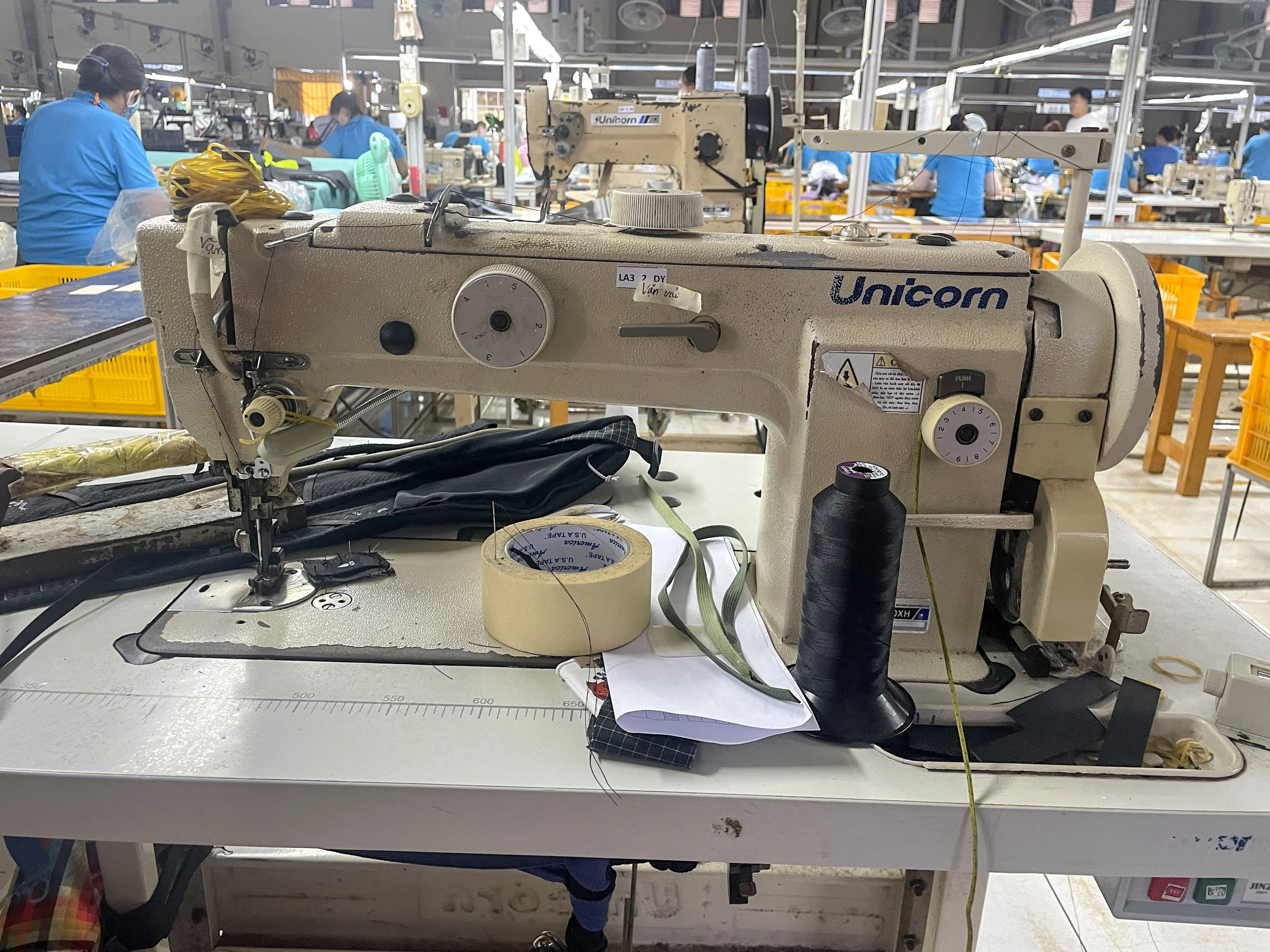 Factory setting with multiple sewing machines and workers in blue shirts working in the background.