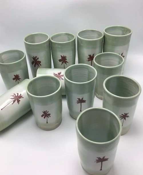 Set of eleven white ceramic cups with brown palm tree designs, arranged on a white surface.
