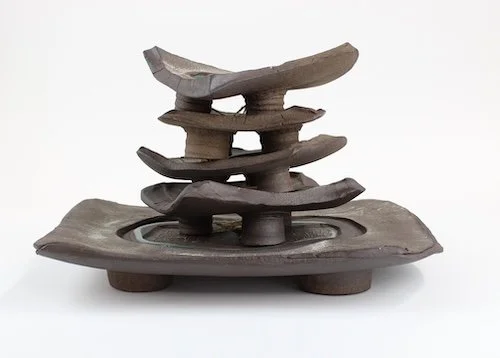 A ceramic sculpture resembling a layered, abstract structure with a rectangular base and multiple stacked, uneven, horizontal plates.