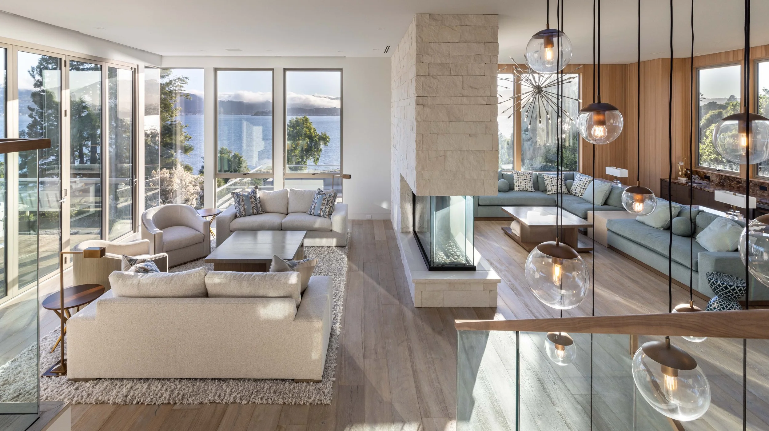 Modern living room with large windows overlooking water and trees, furnished with white and pastel sofas, a stone fireplace, wooden flooring, and hanging glass pendant lights.
