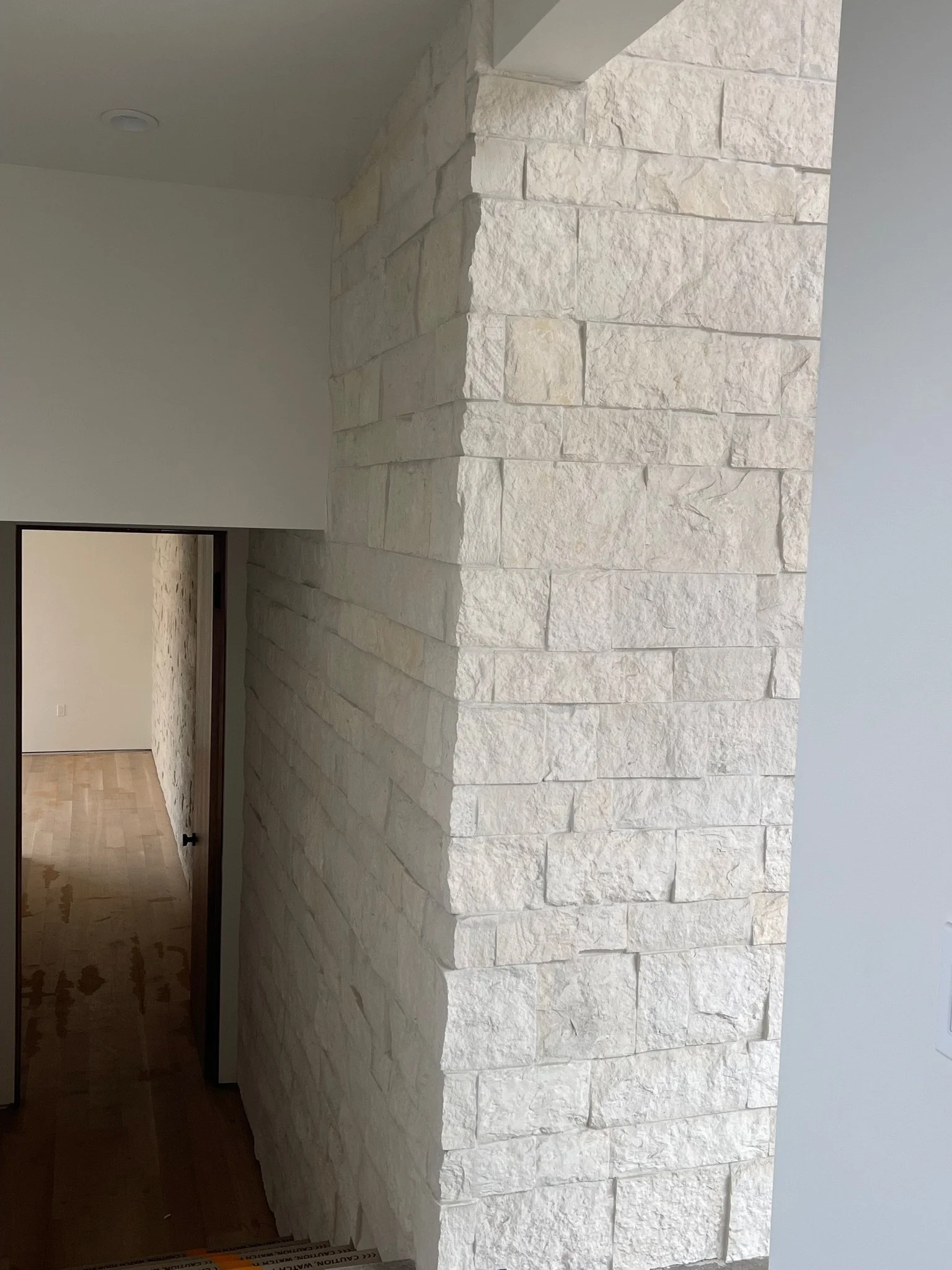 Interior view showing a white stone wall with a stepped pattern next to a white wall and a doorway leading to a room with a wood floor.