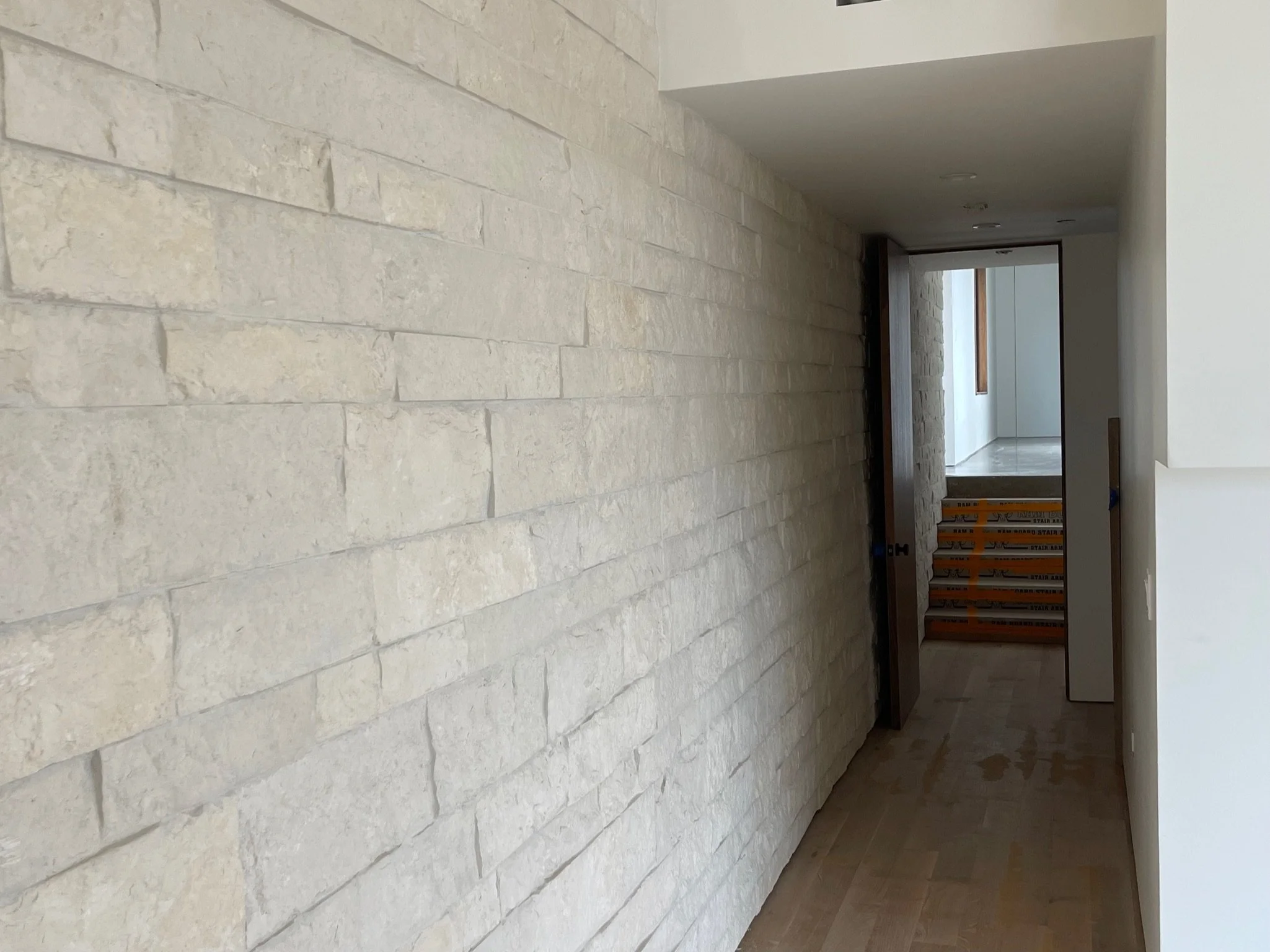 A hallway with a white stone wall on the left and a white wall on the right. At the end of the hallway, there are stairs and a window, with some construction materials visible.