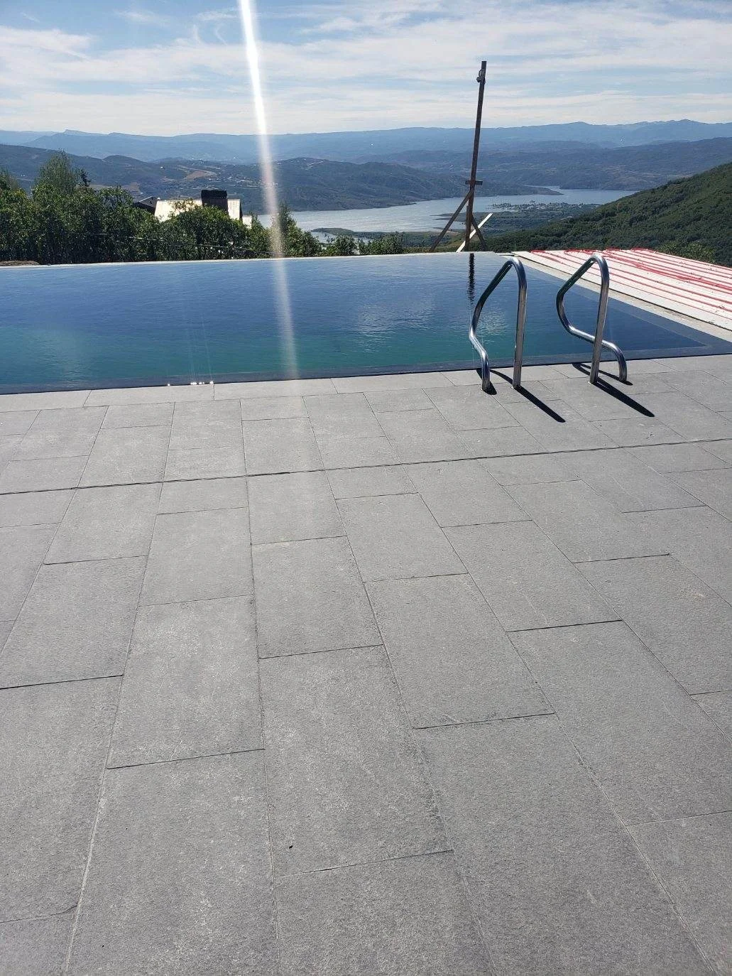 A swimming pool on a tiled deck with an ocean view and mountains in the distance. The pool has a ladder and is surrounded by a scenic landscape.