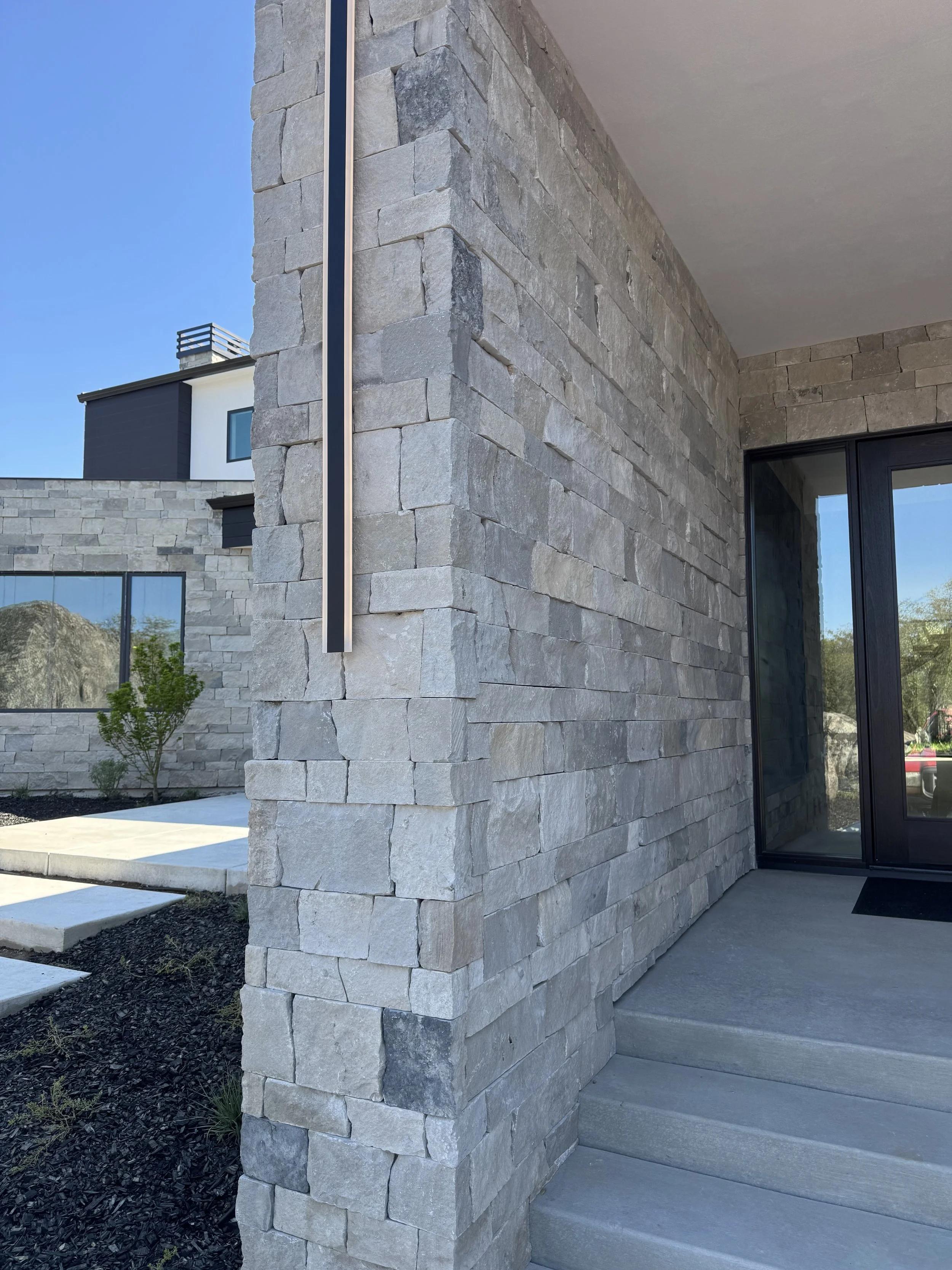Close-up of a modern building exterior showcasing a stone wall with a vertical light fixture, stairs leading to glass doors, and a landscaped area with small plants.