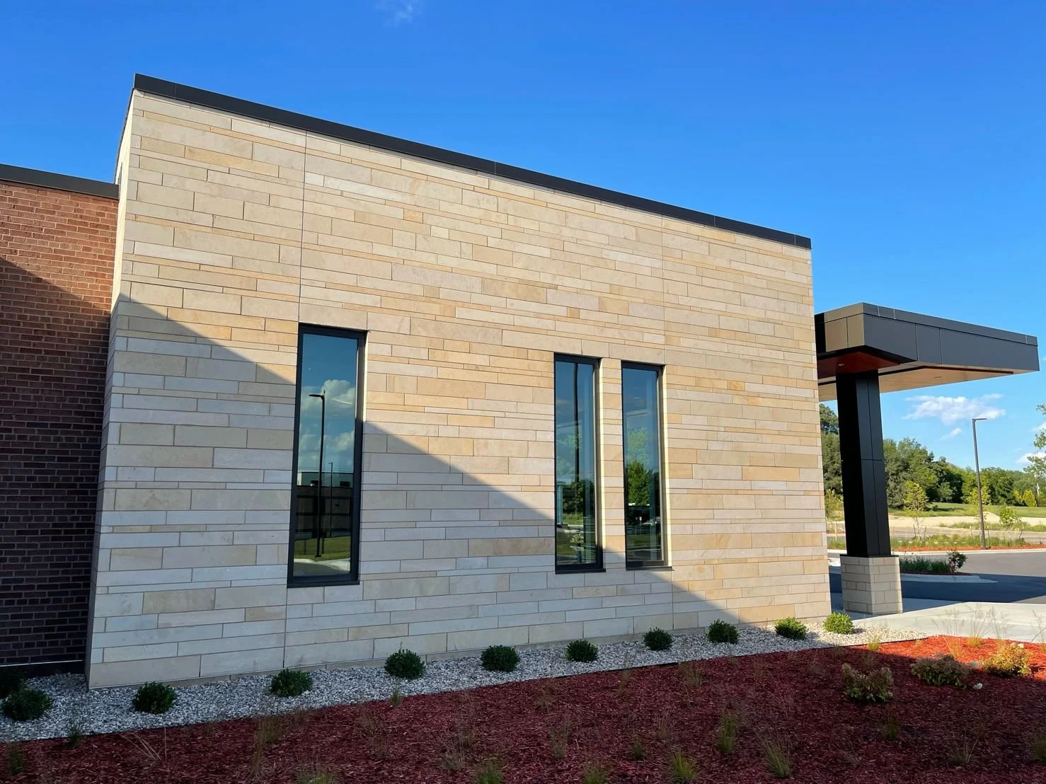 Modern building with beige brick and glass windows, surrounded by landscaped area with small bushes and red mulch, under a clear blue sky.