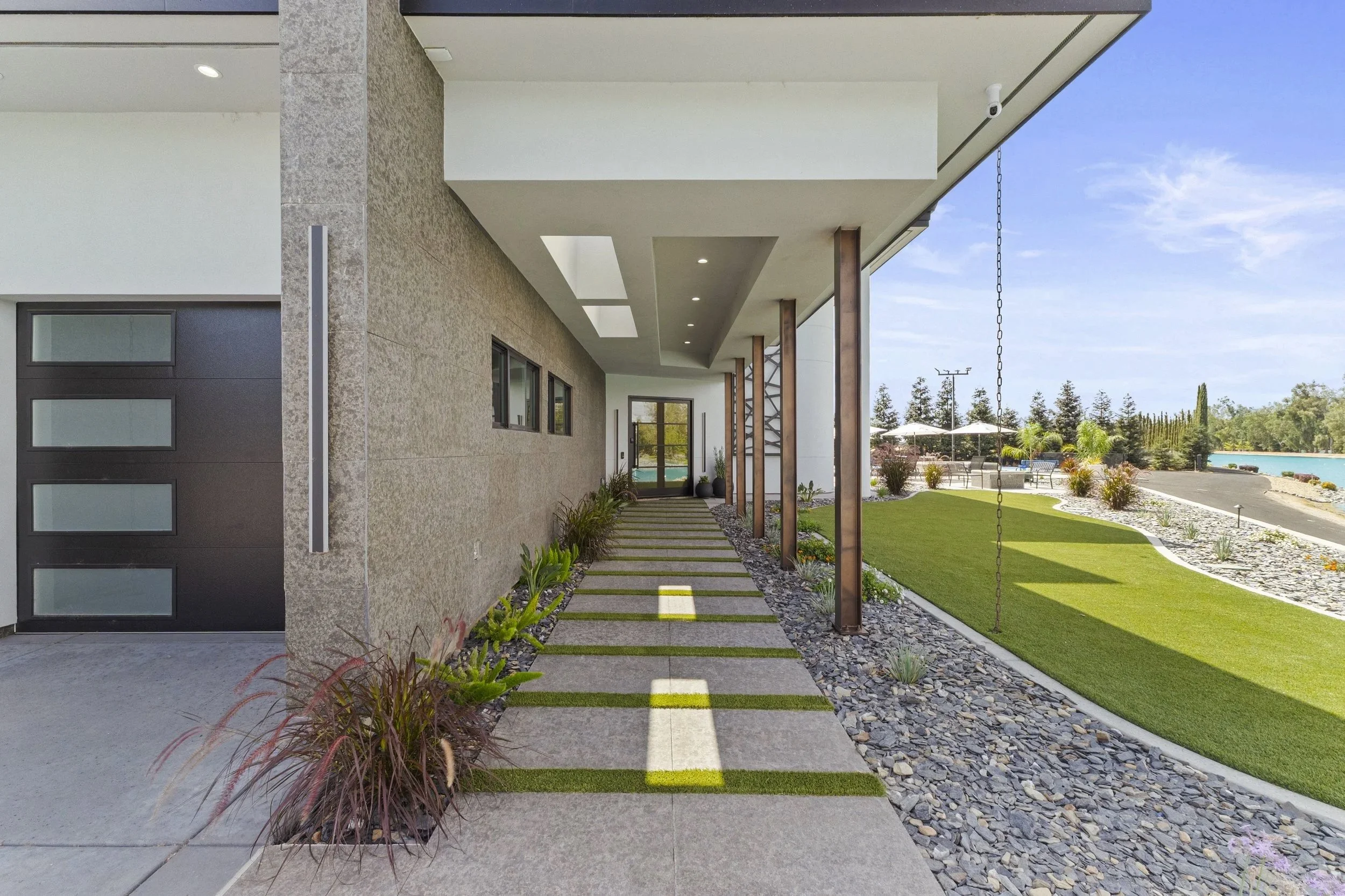 Modern house exterior with a pathway, glass doors, decorative plants, and patio area overlooking a landscaped yard and water in the background.