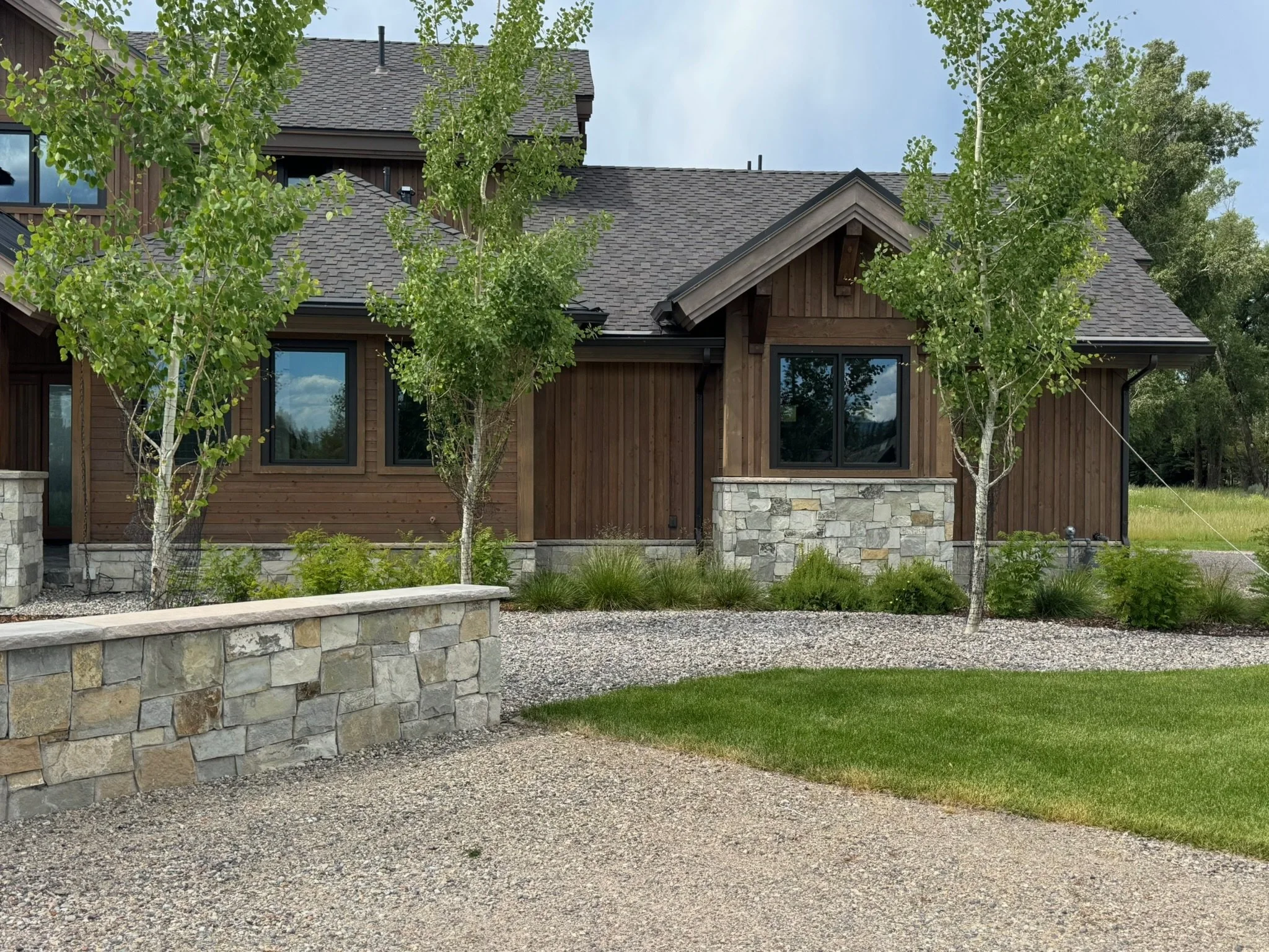 A modern house with wooden exterior walls, stone accents, a gabled roof, surrounded by green trees and landscaping, on a gravel driveway.