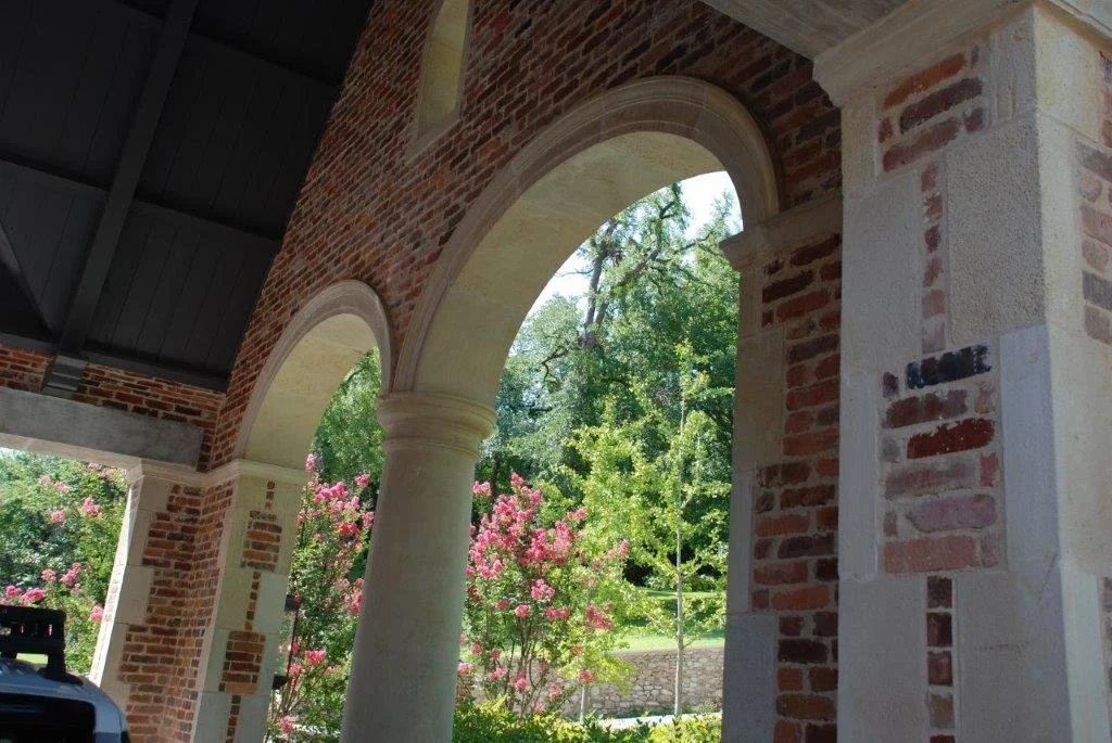 Exterior view of a brick building with stone arches and columns, overlooking a garden with pink flowering bushes and green trees under a sunny sky.