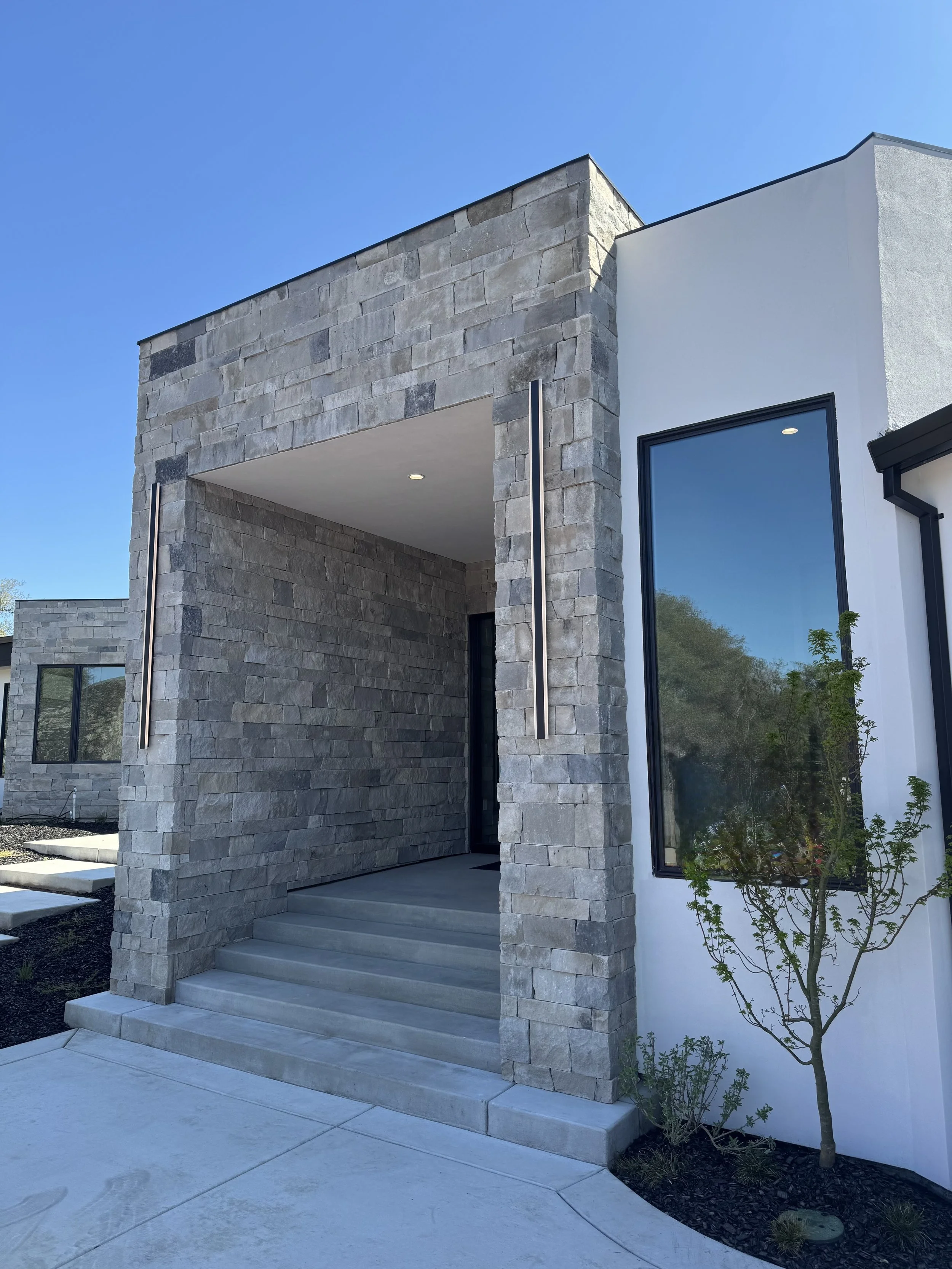 Modern house exterior with a stone facade, wide glass windows, and a small tree near the entrance under clear blue sky.