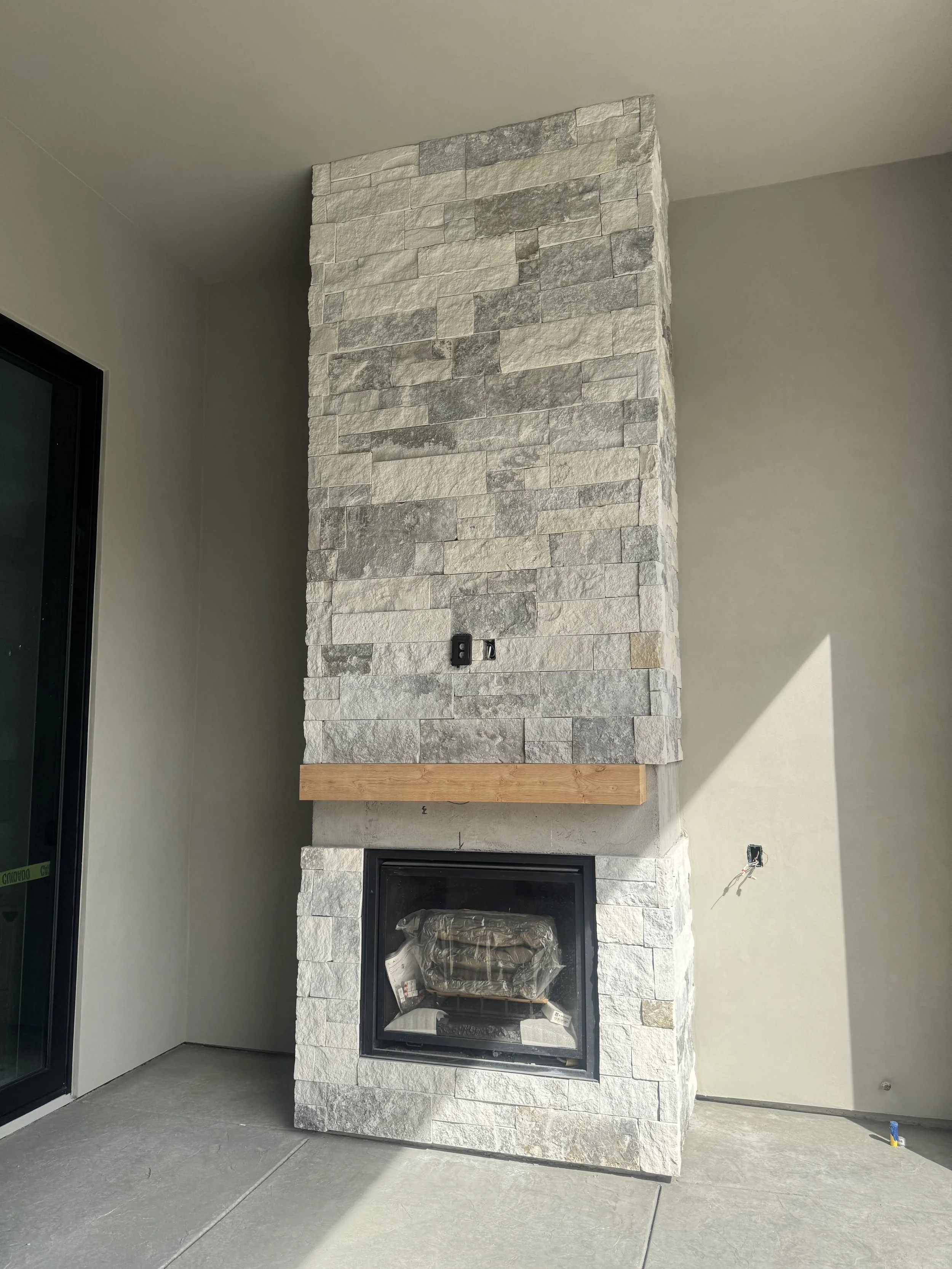 Stone fireplace under construction with a black glass front, a wooden mantel, and electrical outlets, next to a sliding glass door in a modern home.