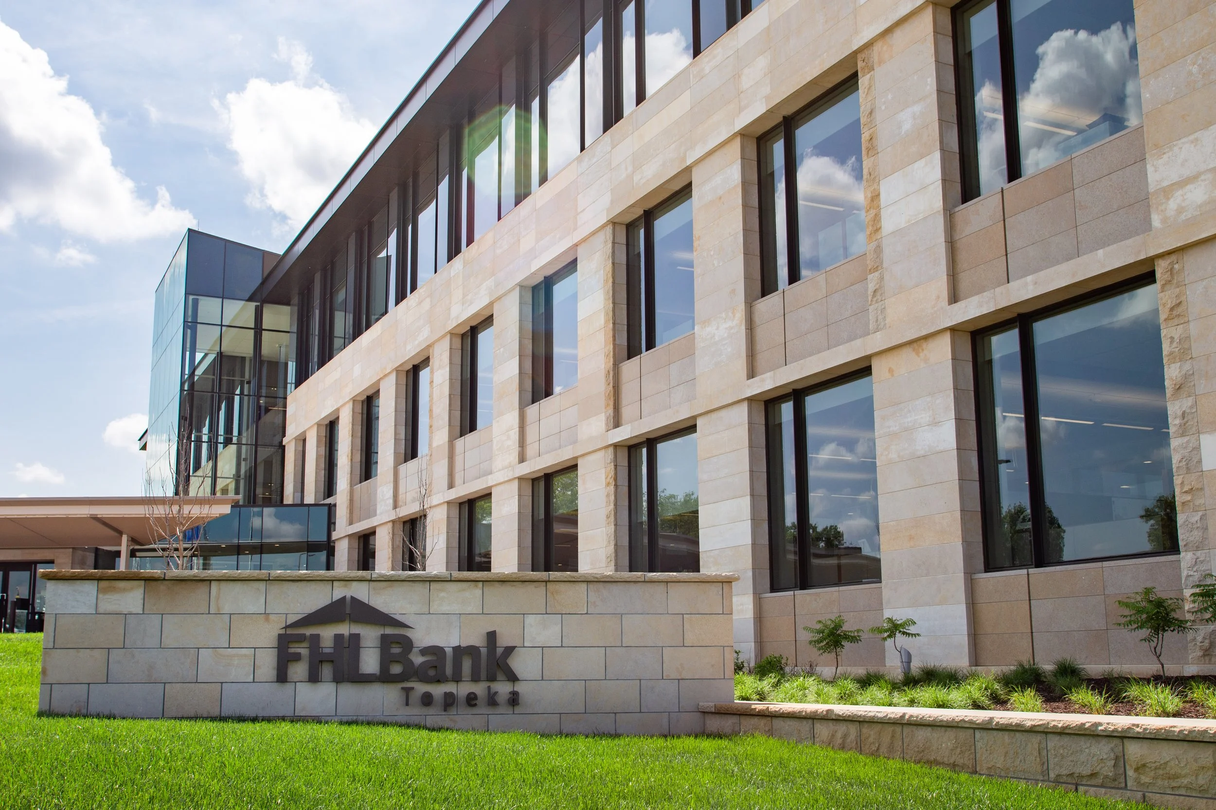 Modern commercial building with reflective windows and a stone facade, signage reading 'FHL Bank Topeka' in front of a well-maintained lawn under a partly cloudy sky.