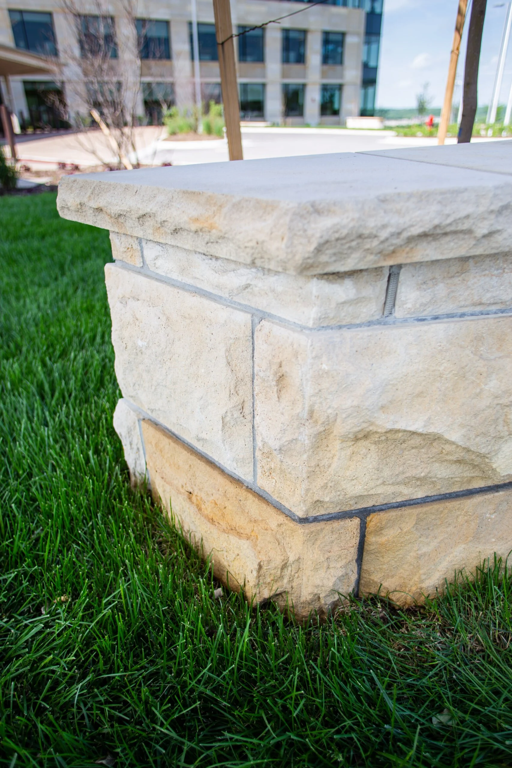 Close-up of a stone outdoor planter with a concrete top, situated on green grass with a modern building in the background.