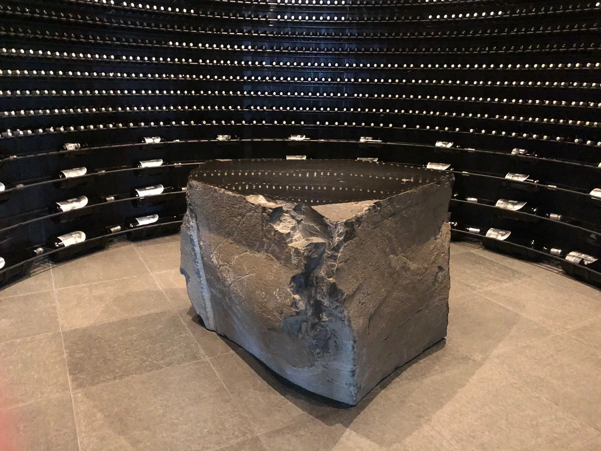 A large rough stone block in a wine cellar, surrounded by horizontal racks of wine bottles stored on metal racks.