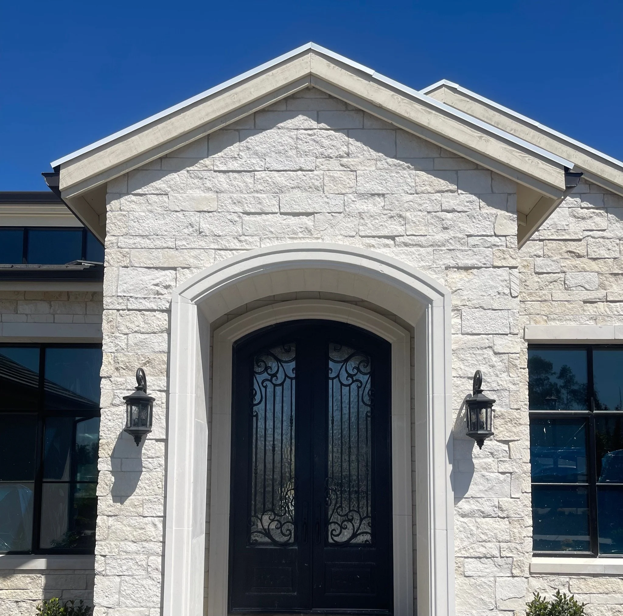 Front view of a house with a stone facade, black iron door with ornate design, and two wall-mounted lanterns, under a blue sky.
