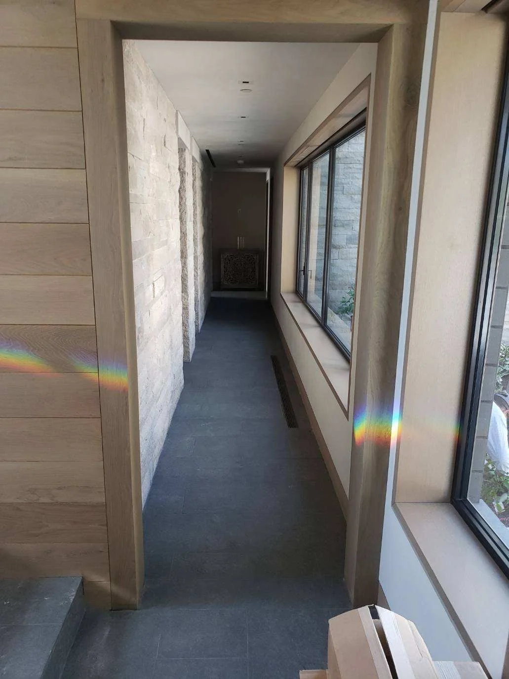 A narrow hallway with a stone wall on the left and large windows on the right, showing an outdoor view. Sunlight creates rainbow reflections on the wall and window.
