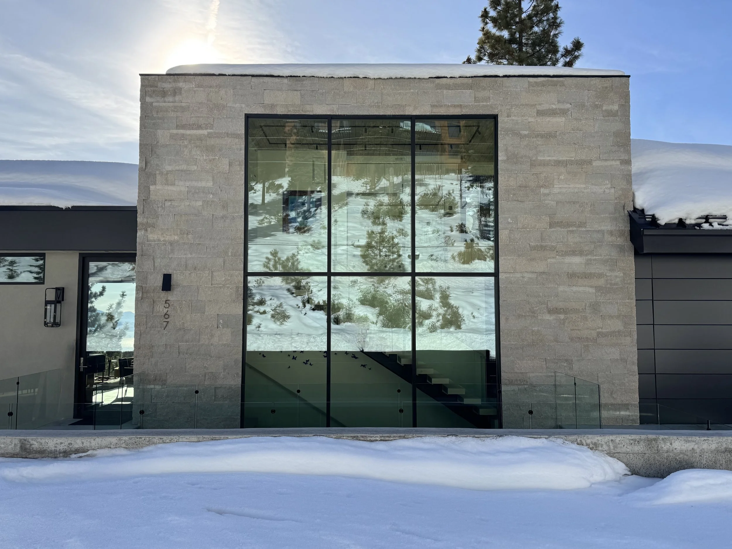 Modern house with large glass window, stone exterior, snow on the ground and roof, trees reflected in the window, and a partly cloudy sky.