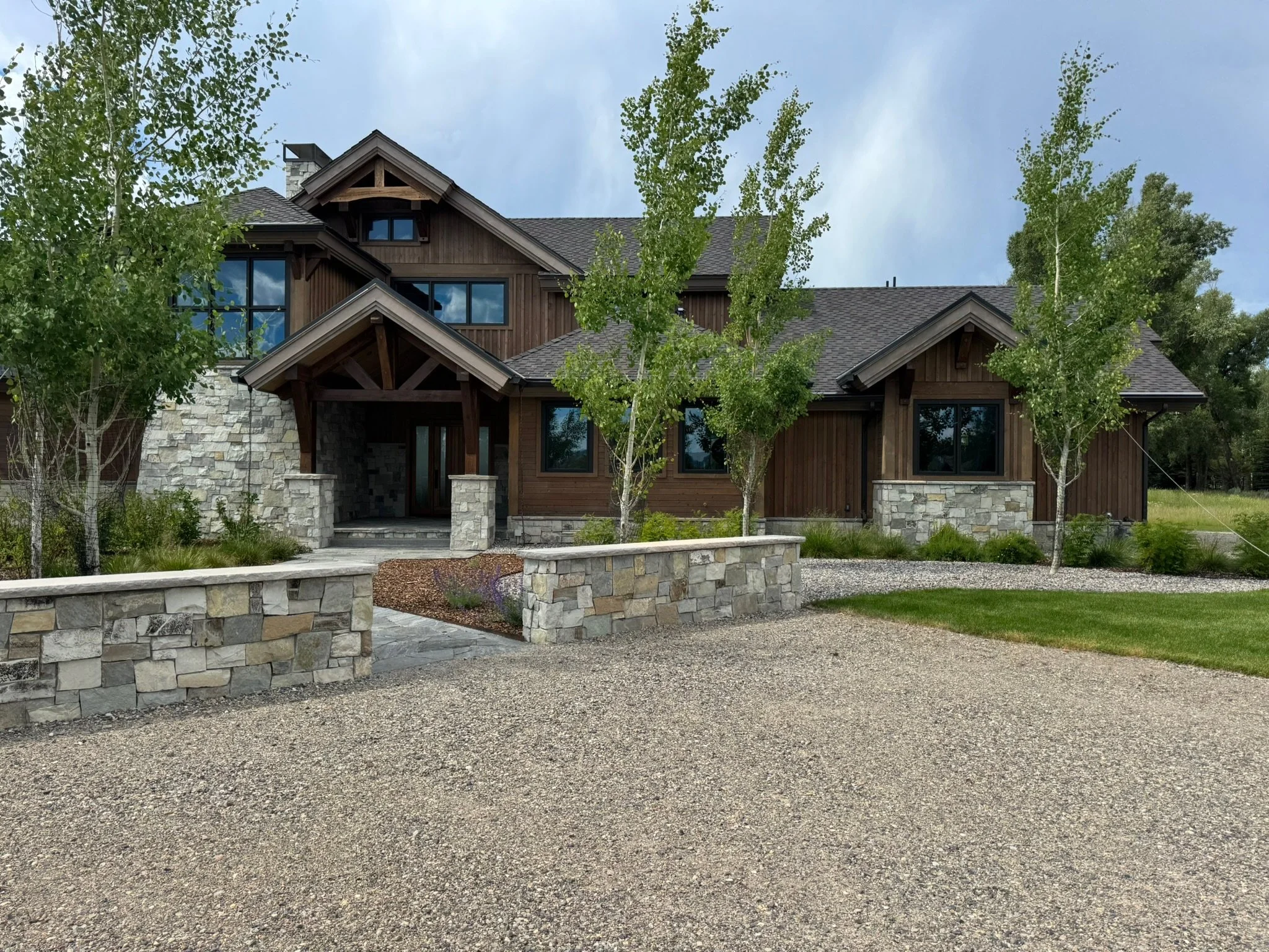 Large modern house with a mix of wood and stone exterior, surrounded by trees and landscaped yard with a gravel driveway.