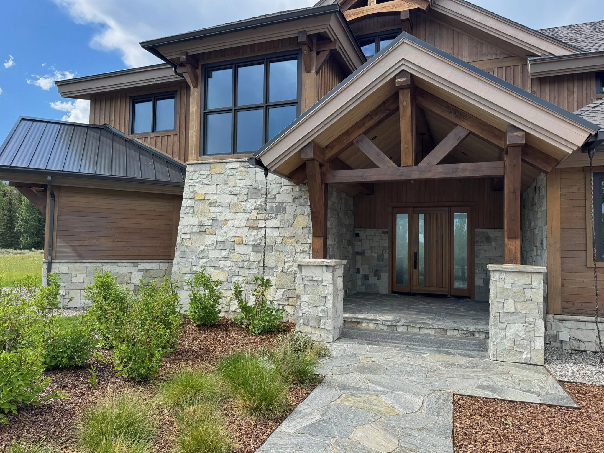 Front entrance of a house with stone and wood exterior, glass double doors, covered porch, stone pathway, and garden plants.
