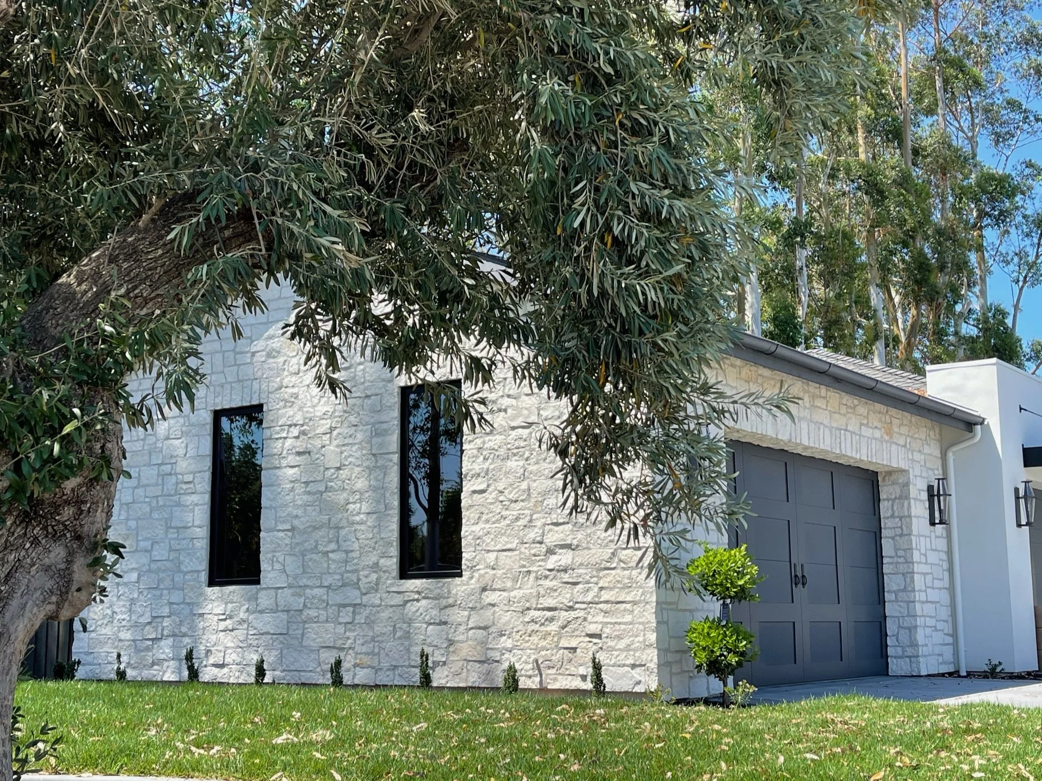 Modern white house with a gray garage door, surrounded by green grass, small trees, and tall trees in the background under a blue sky.