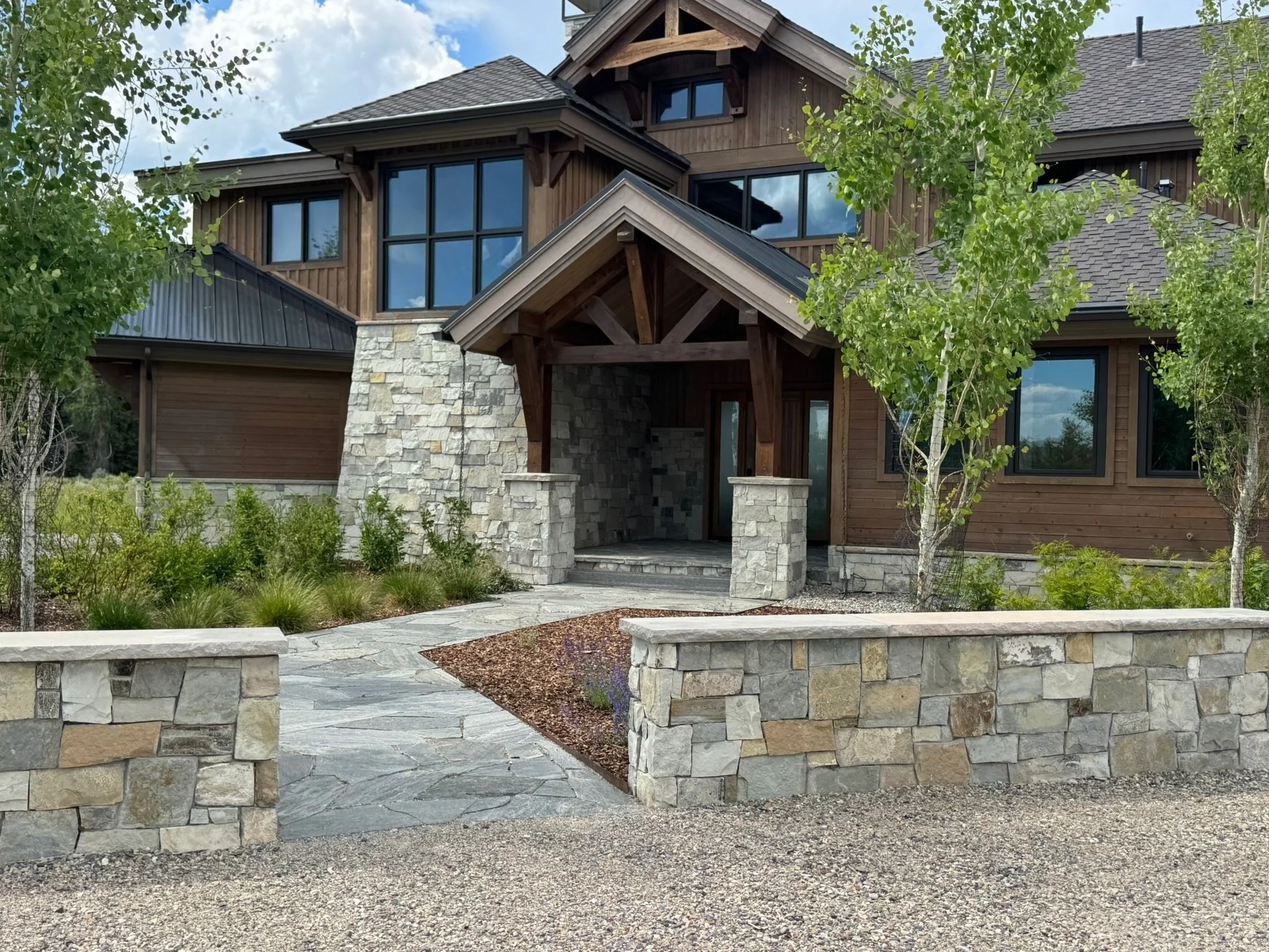 Modern house with a stone and wood exterior, large windows, a small stone pathway, and trees in front.