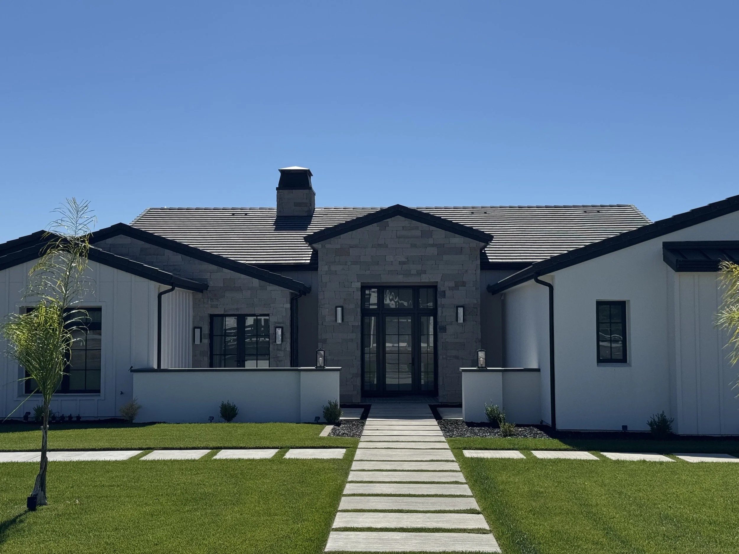Modern house with a stone and white siding exterior, black window frames, a pathway leading to the front door, and a well-maintained lawn with small trees and shrubs, under a clear blue sky.