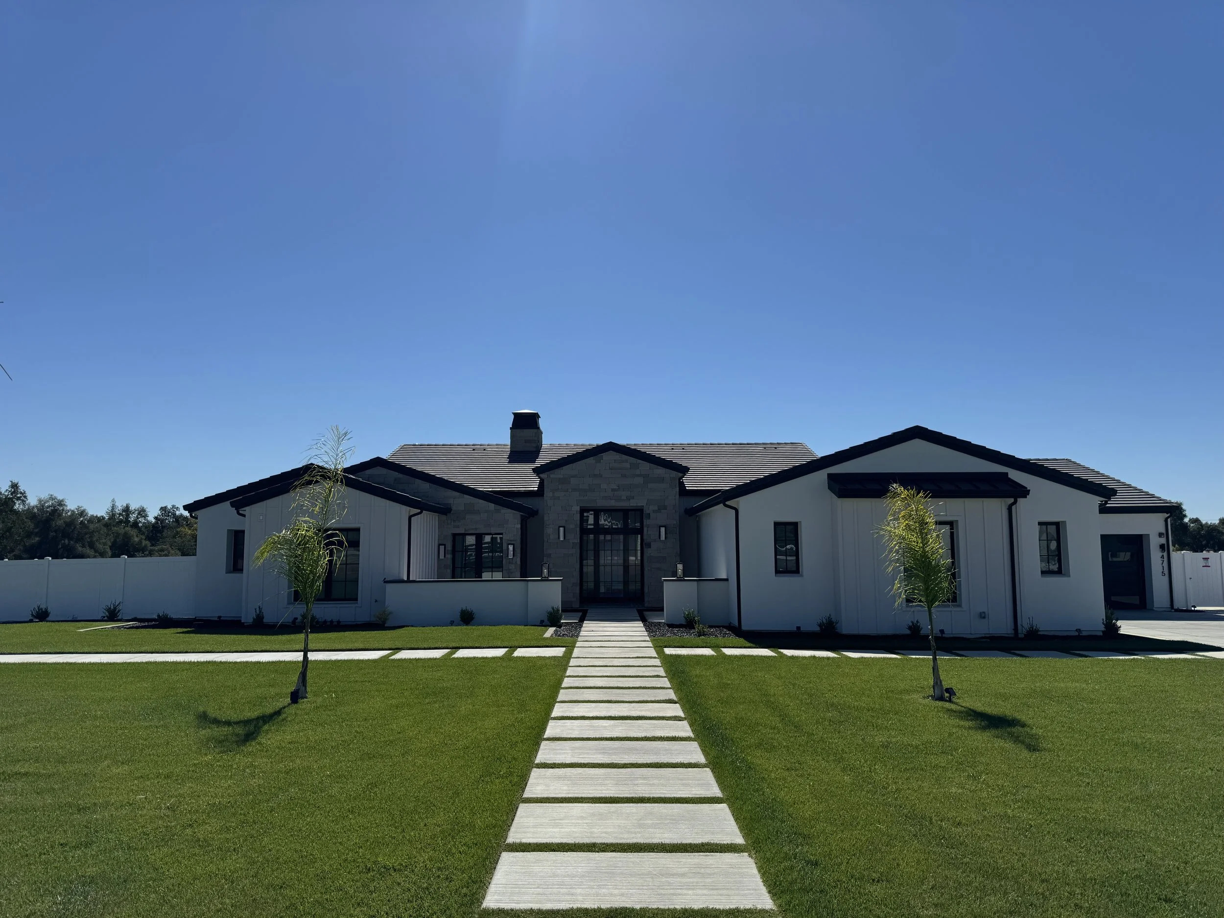Modern house with white and gray exterior, black roof, with a walkway leading to the front door, landscaped lawn with small trees, under a clear blue sky.