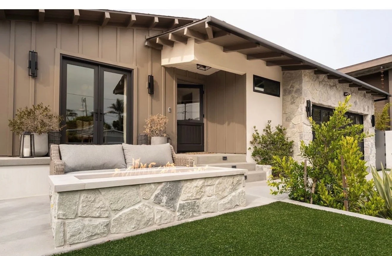 Rear view of a modern house with a patio, stone and wood exterior, sliding glass doors, a stone fire pit with cushions, plants, and a lawn.