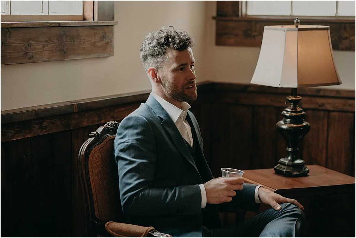 groom in blue suit sitting down and drinking in a rustic room