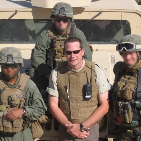 Group of four soldiers in military gear with one civilian in front, standing in front of a military vehicle.