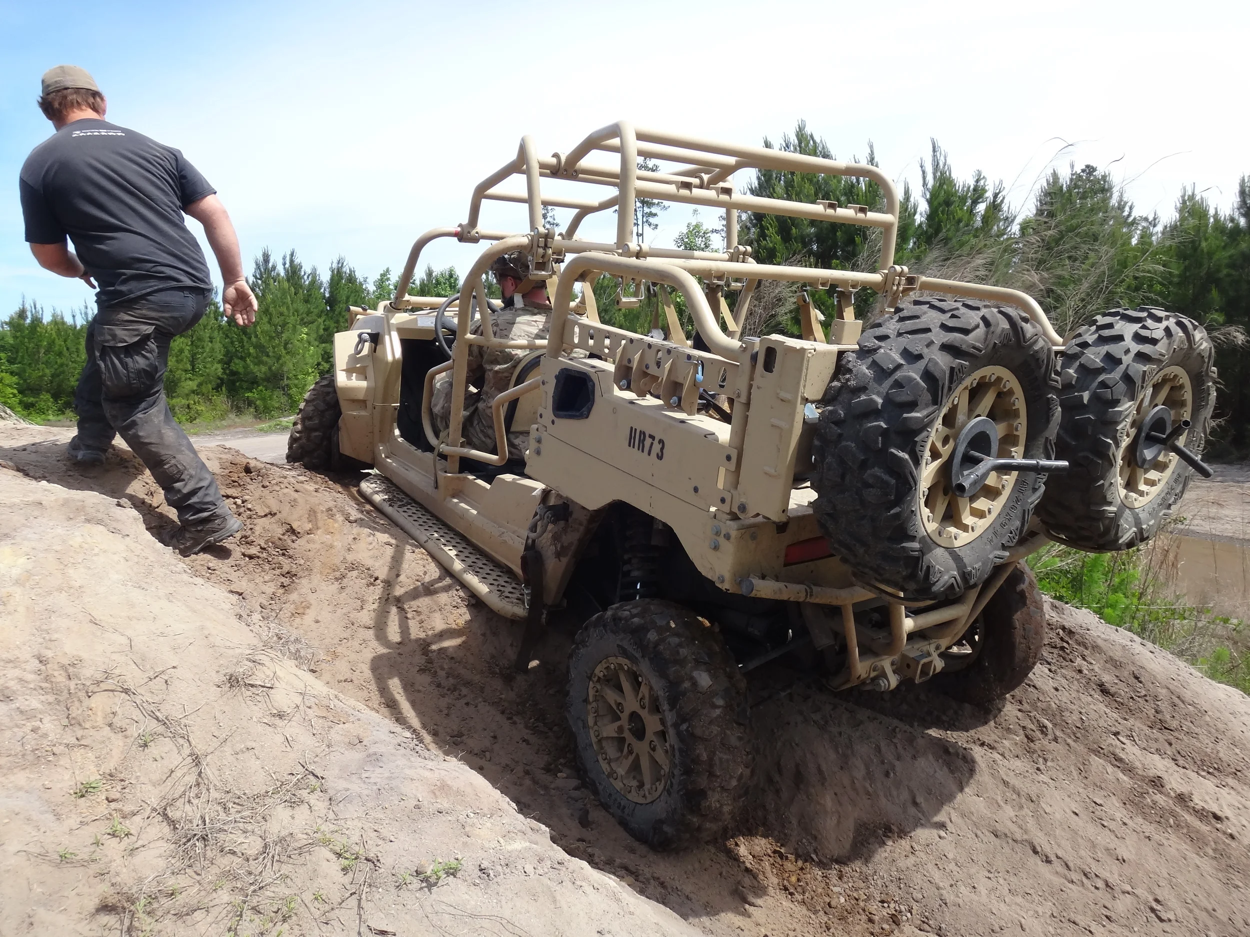 Armored military vehicle stuck in a deep dirt rut during military training and mobility training, with a man in a black T-shirt and cargo pants walking beside it under a clear blue sky with green trees.