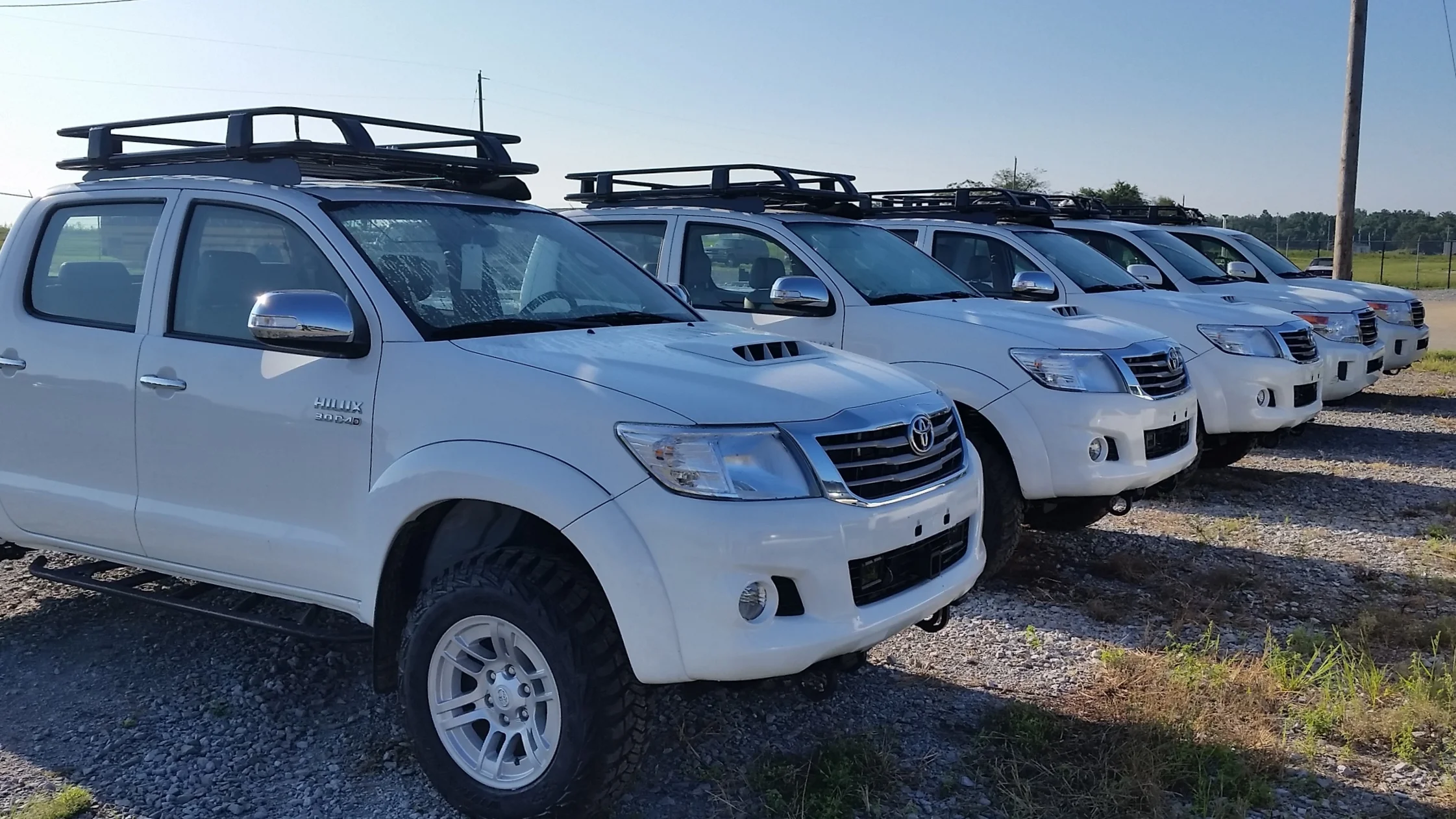 Row of white pickup trucks parked outdoors on a gravel lot, each equipped with a roof rack for military training and mobility training.
