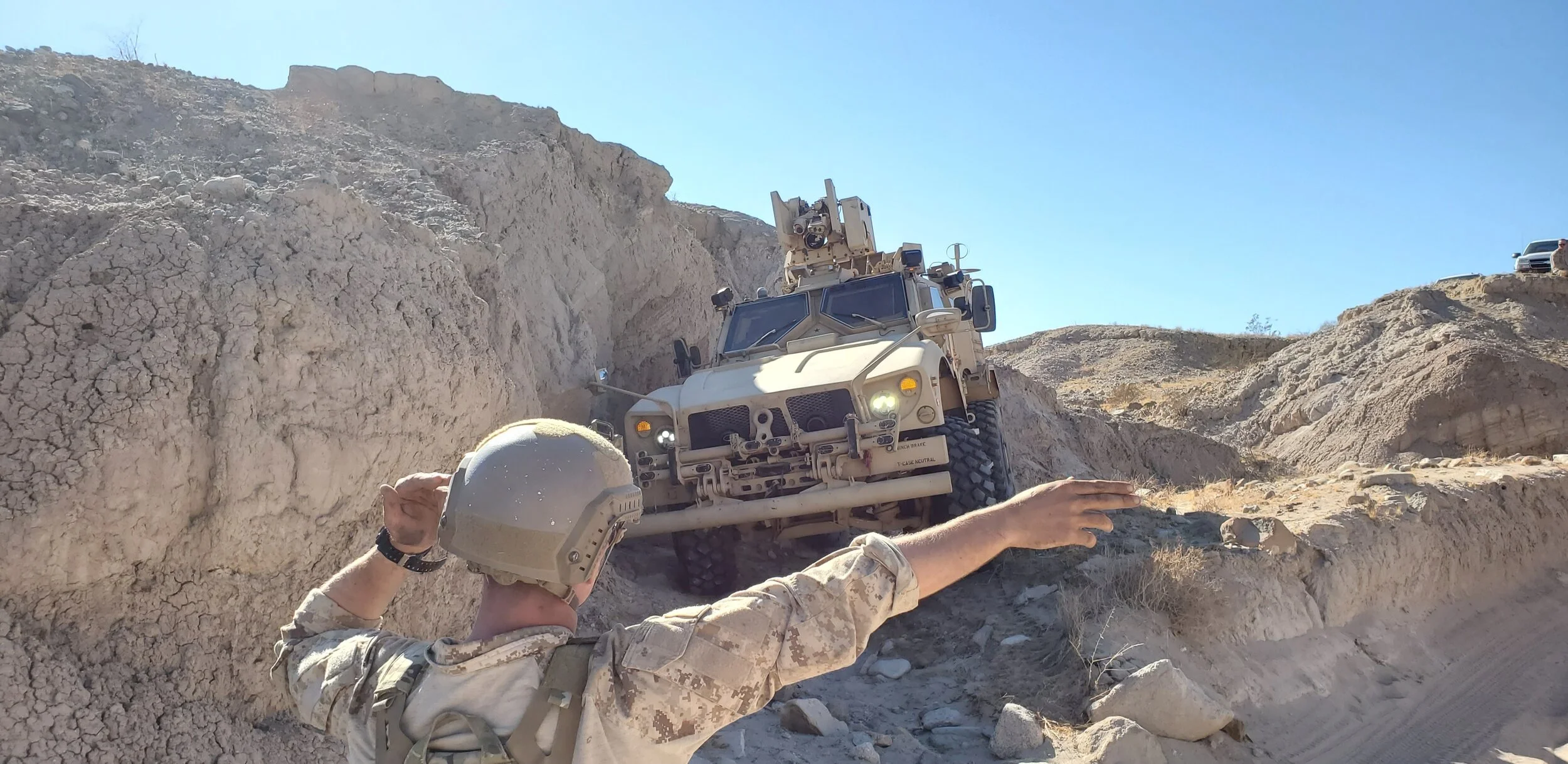 A person in military uniform directing a military vehicle up a rocky dirt slope in a desert environment under a clear blue sky.