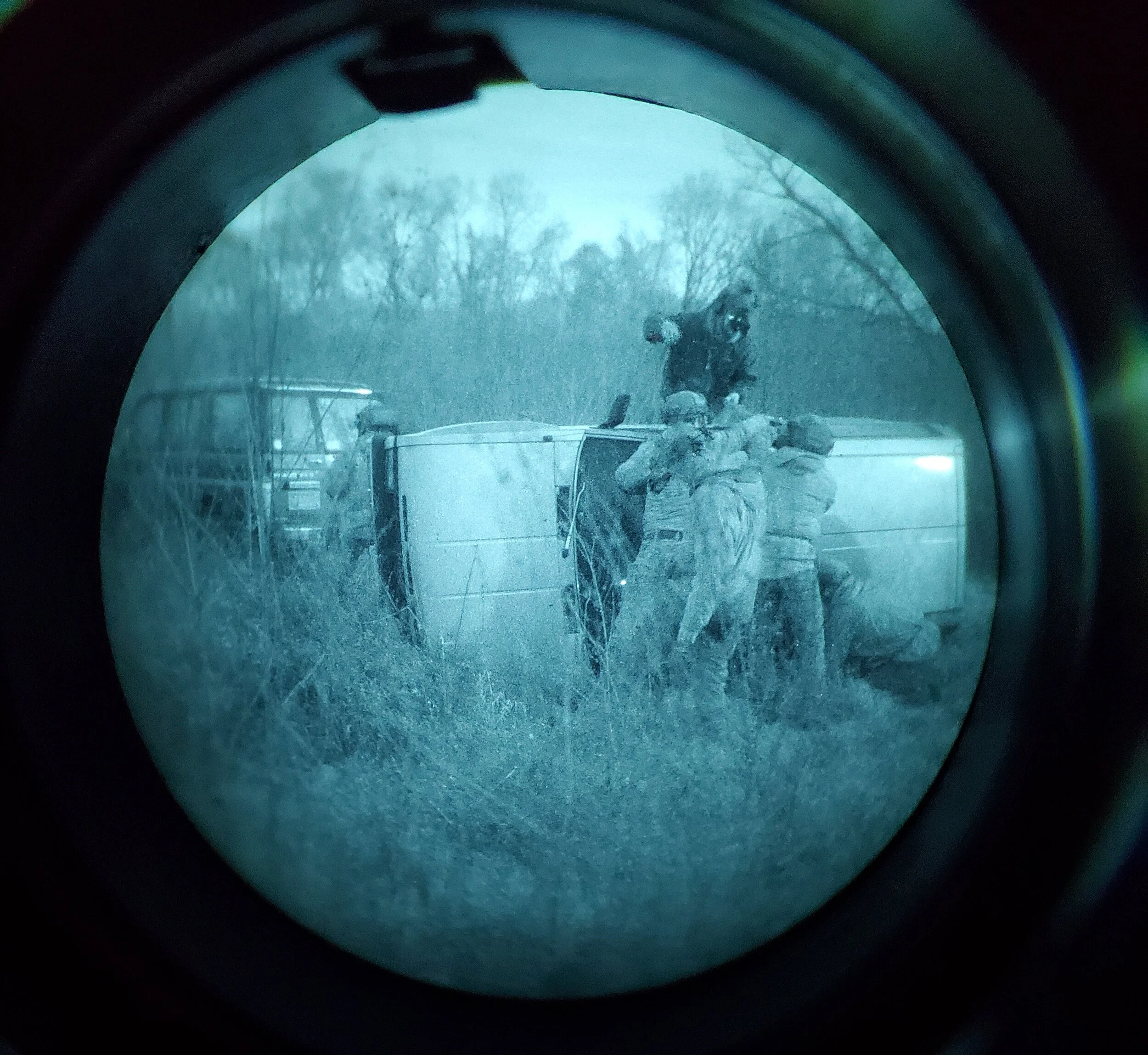 View through a rifle scope showing a person in camouflage clothing crouching behind a fallen log in a wooded area during winter mobility training. The scene is foggy with bare trees, and the scope's crosshairs are visible in the center.