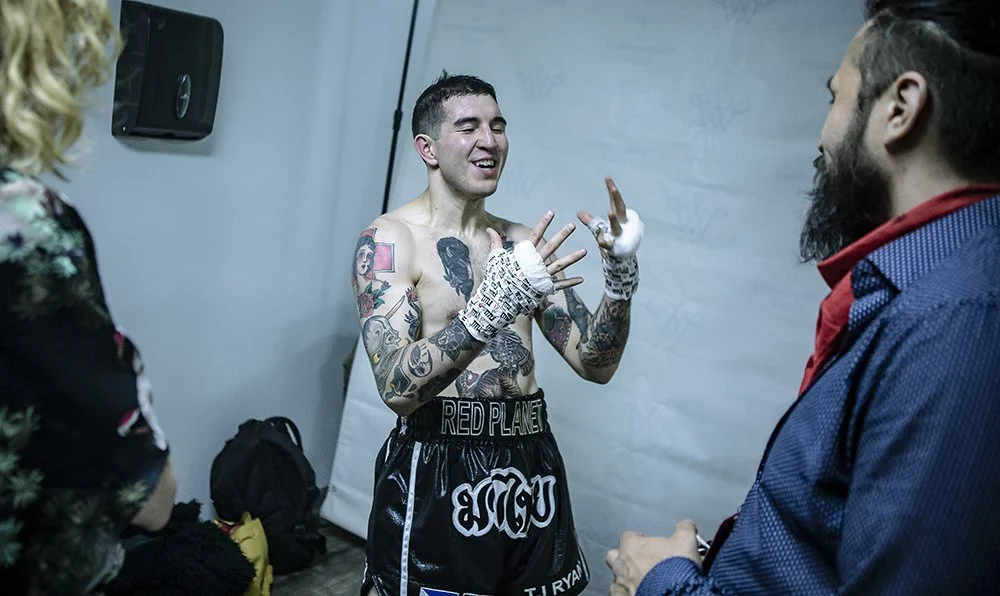 A tattoos boxer with hand wraps smiling and talking to two people in a dressing room or gym.
