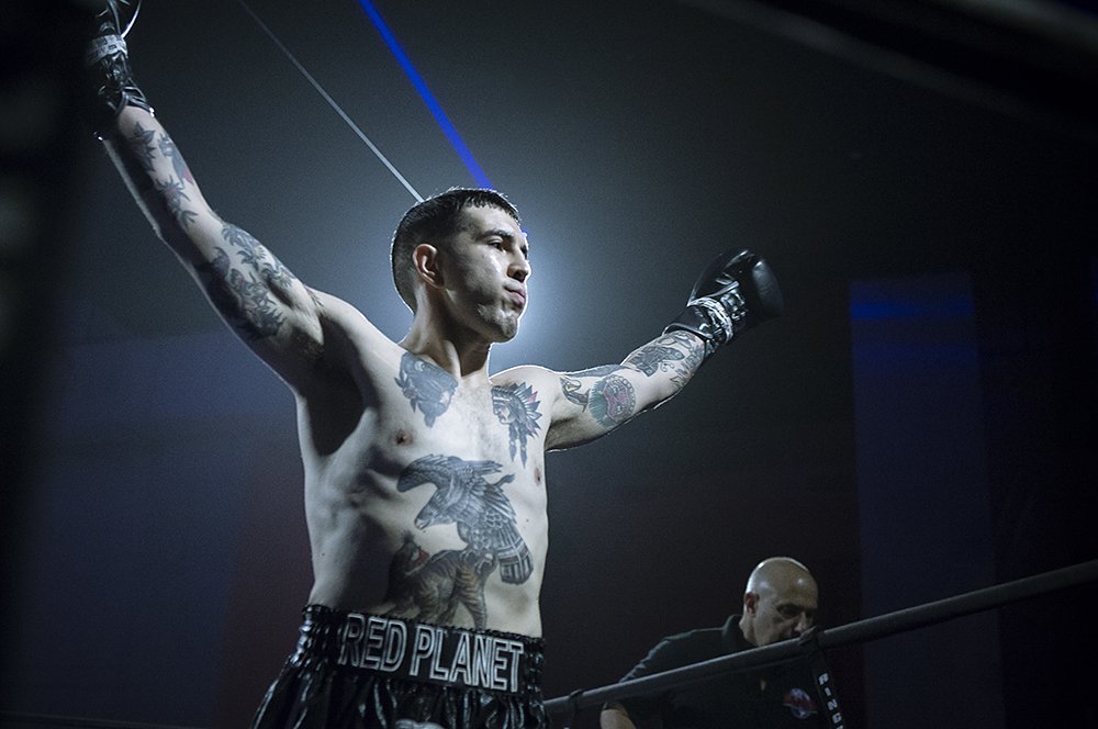 A male boxer with tattoos on his chest and arms raising his arms in a boxing ring, wearing black gloves and shorts with "Red Planet" written on the waistband, with spotlights and a referee in the background.