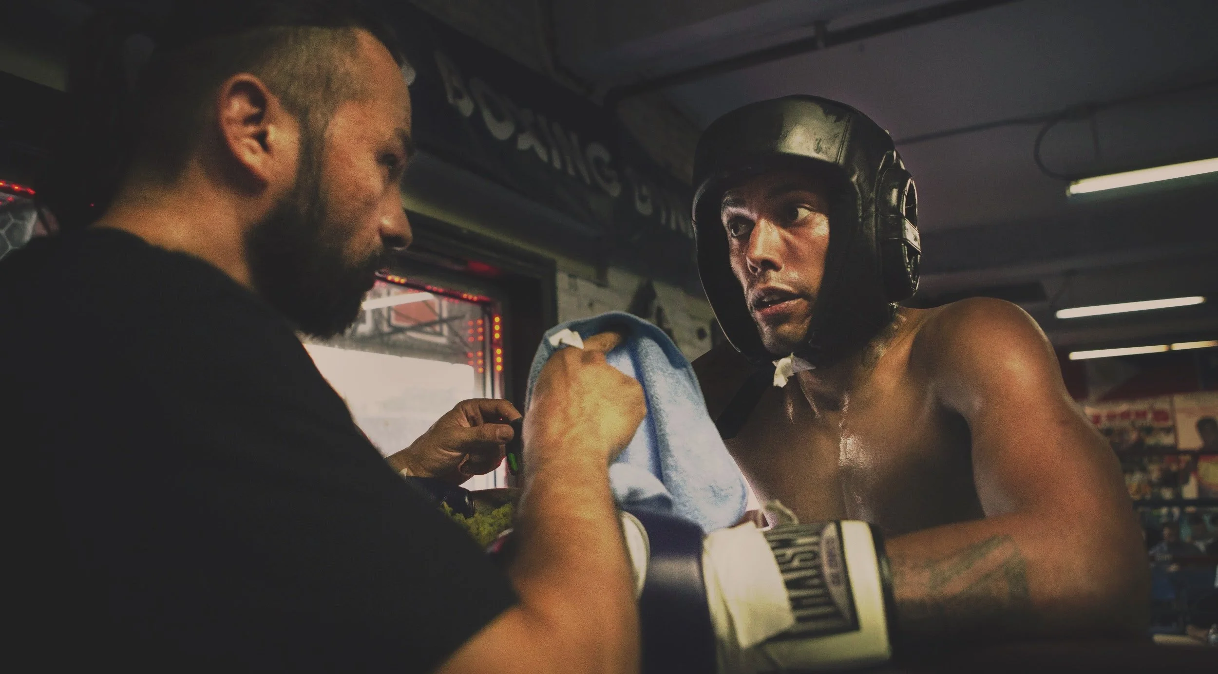 A shirtless boxer with boxing gloves receives medical attention from a man holding a towel, inside a boxing gym.