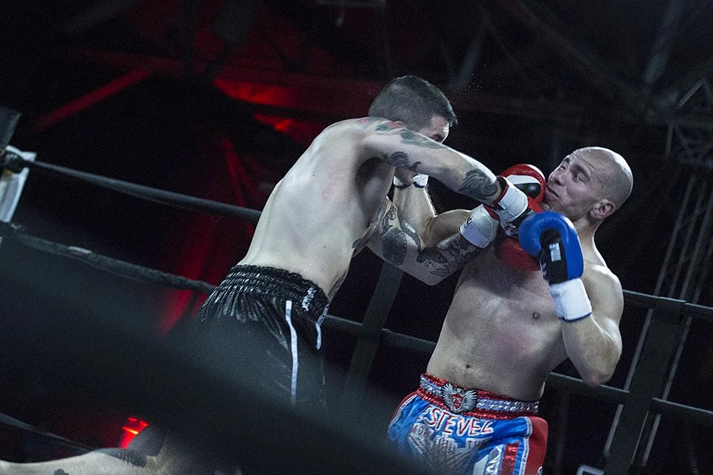 Two male boxers sparring in a boxing ring, one delivering a punch to the other's face, with a dark background and red and blue lighting.