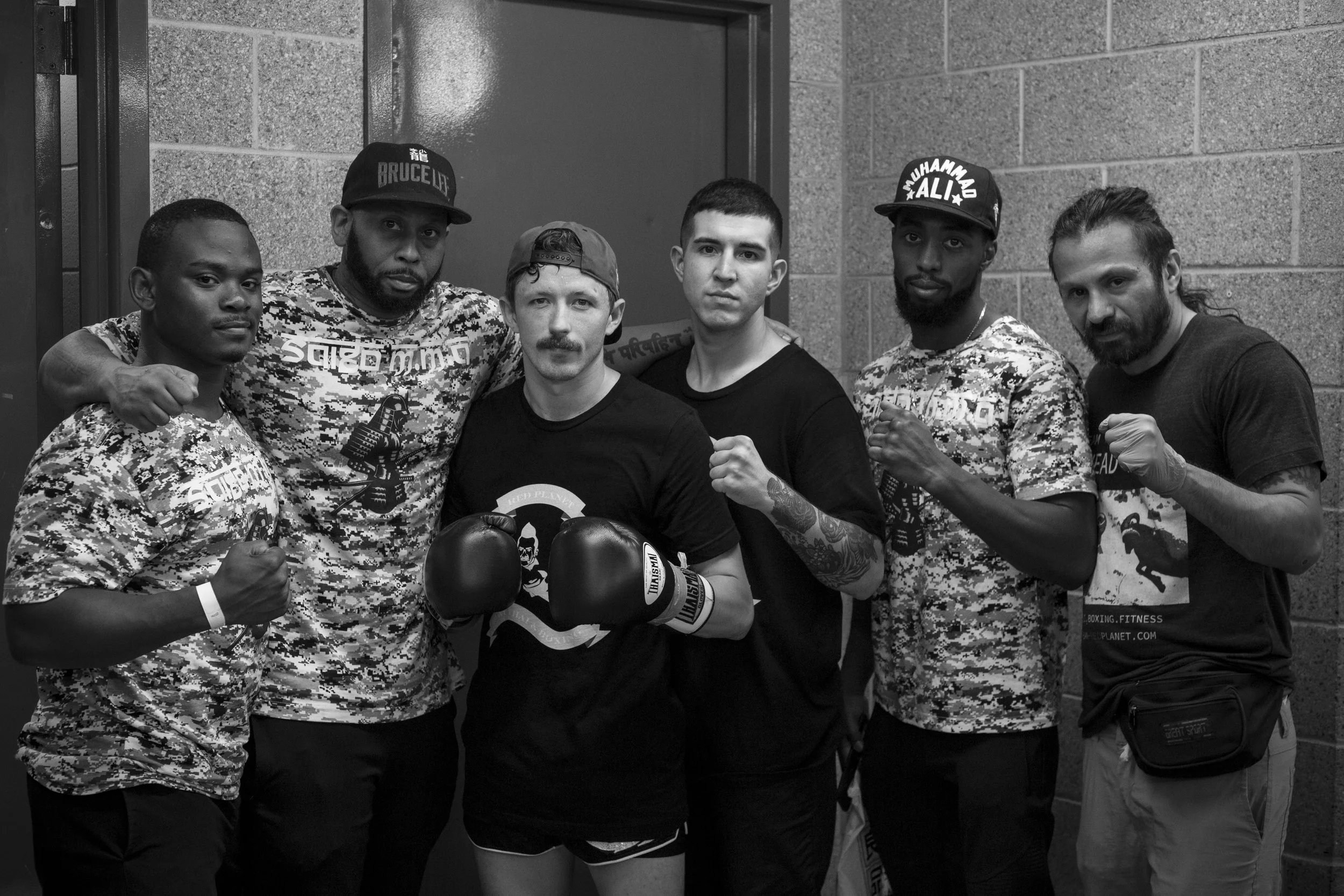 Six men posing together, some in boxing stance with fists raised, in an indoor setting with brick and concrete walls.