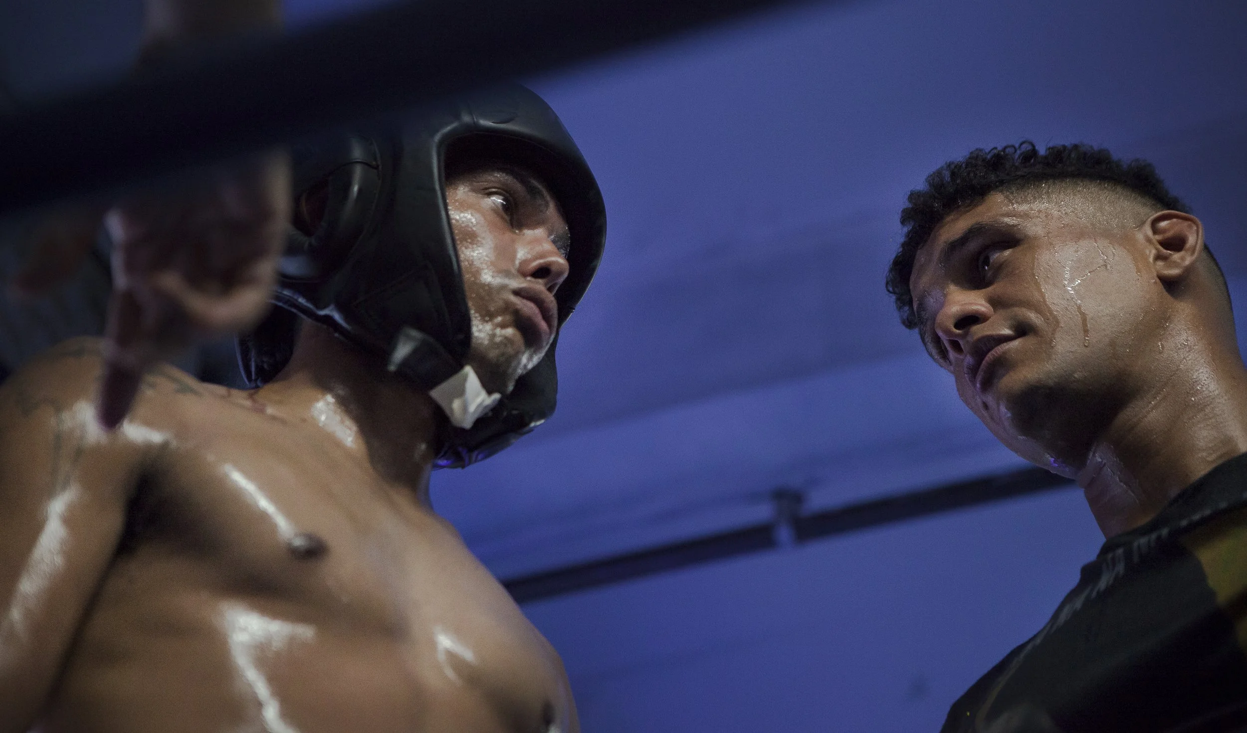 Two shirtless men in a boxing gym, one wearing a black helmet, are having a serious conversation, with sweat visible on their faces.