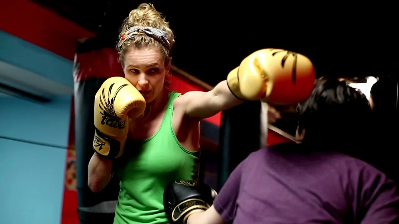 A woman in a green shirt boxing with a trainer in a purple shirt at a gym, woman wearing yellow boxing gloves, woman throwing a punch.
