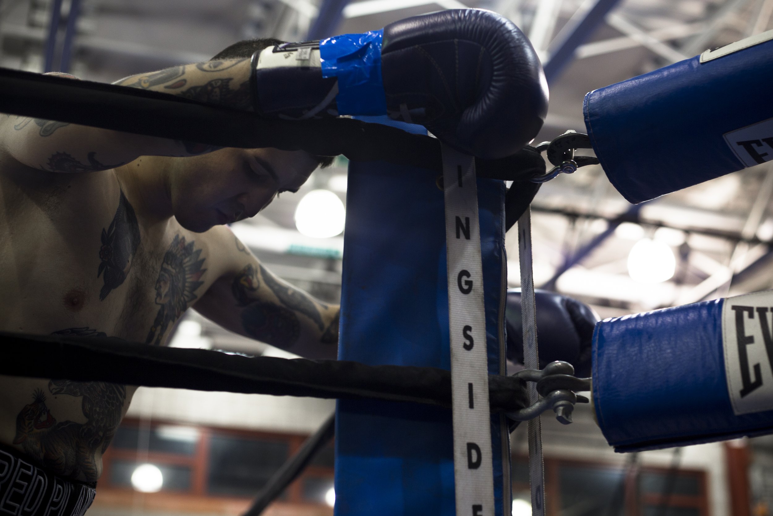 A male boxer with tattoos resting against the corner of a boxing ring, wearing boxing gloves and shorts.
