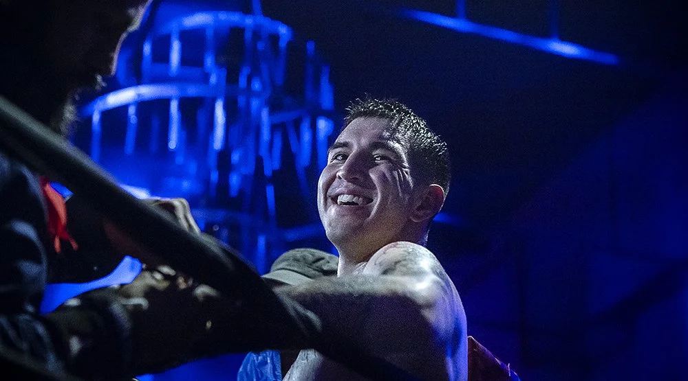 A man smiling and sweating during a boxing match, with a blue-lit background.