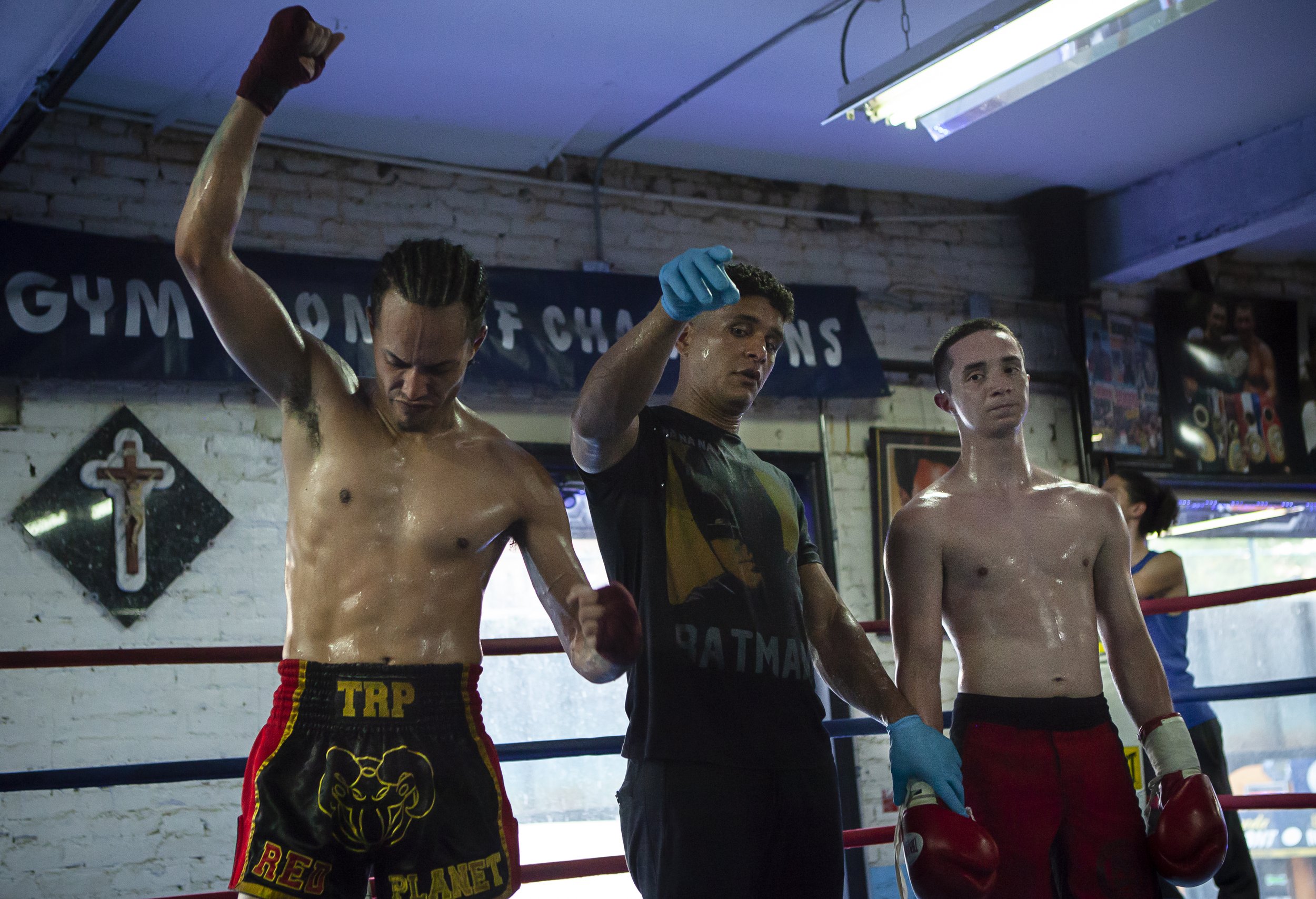 Two shirtless male boxers wearing red shorts with gloves, and a referee in a boxing gym, in front of a banner that reads 'GYM OF CHAMPIONS'.
