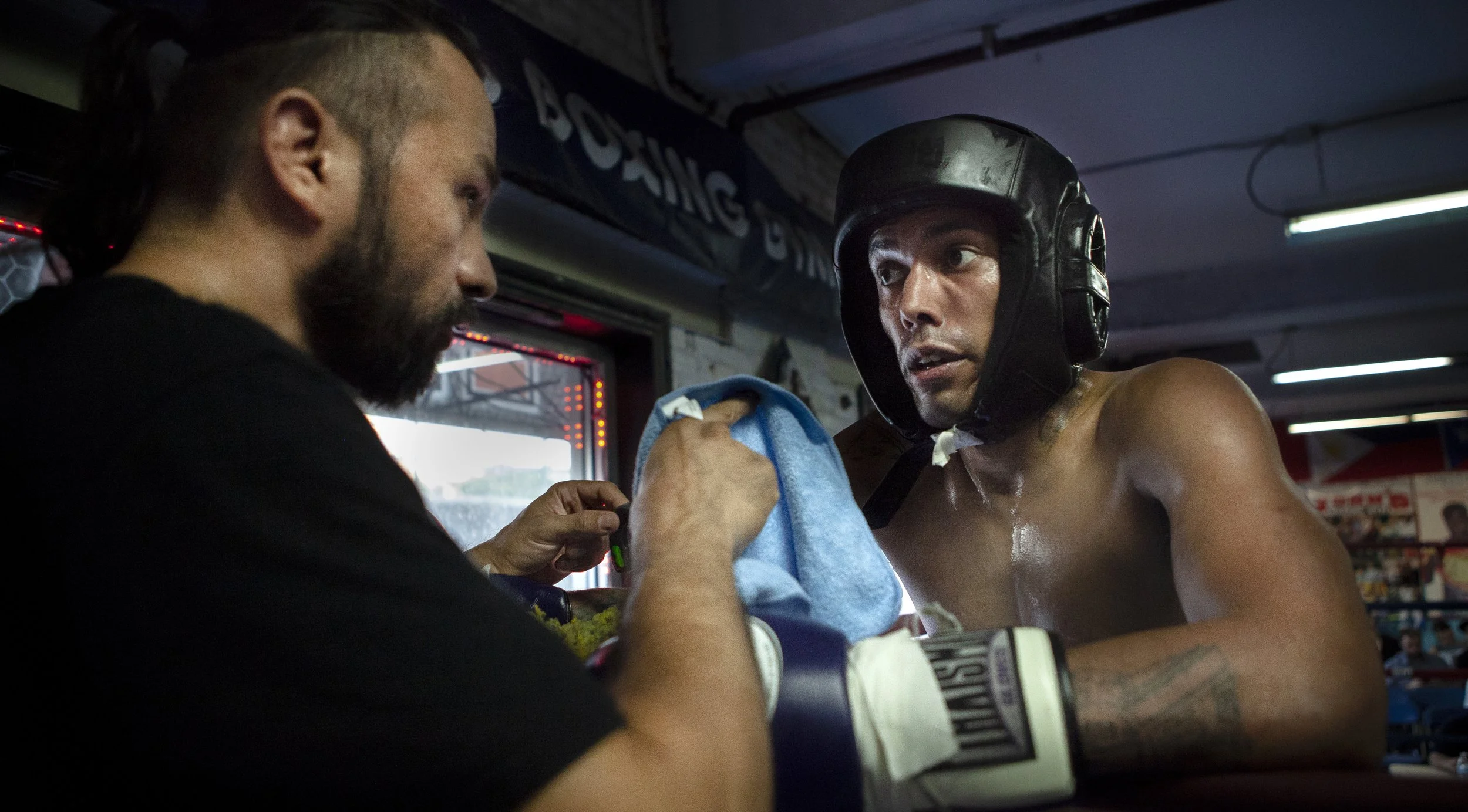A shirtless boxing man wearing a black headgear getting a towel from a man with a beard in a gym or boxing training facility.