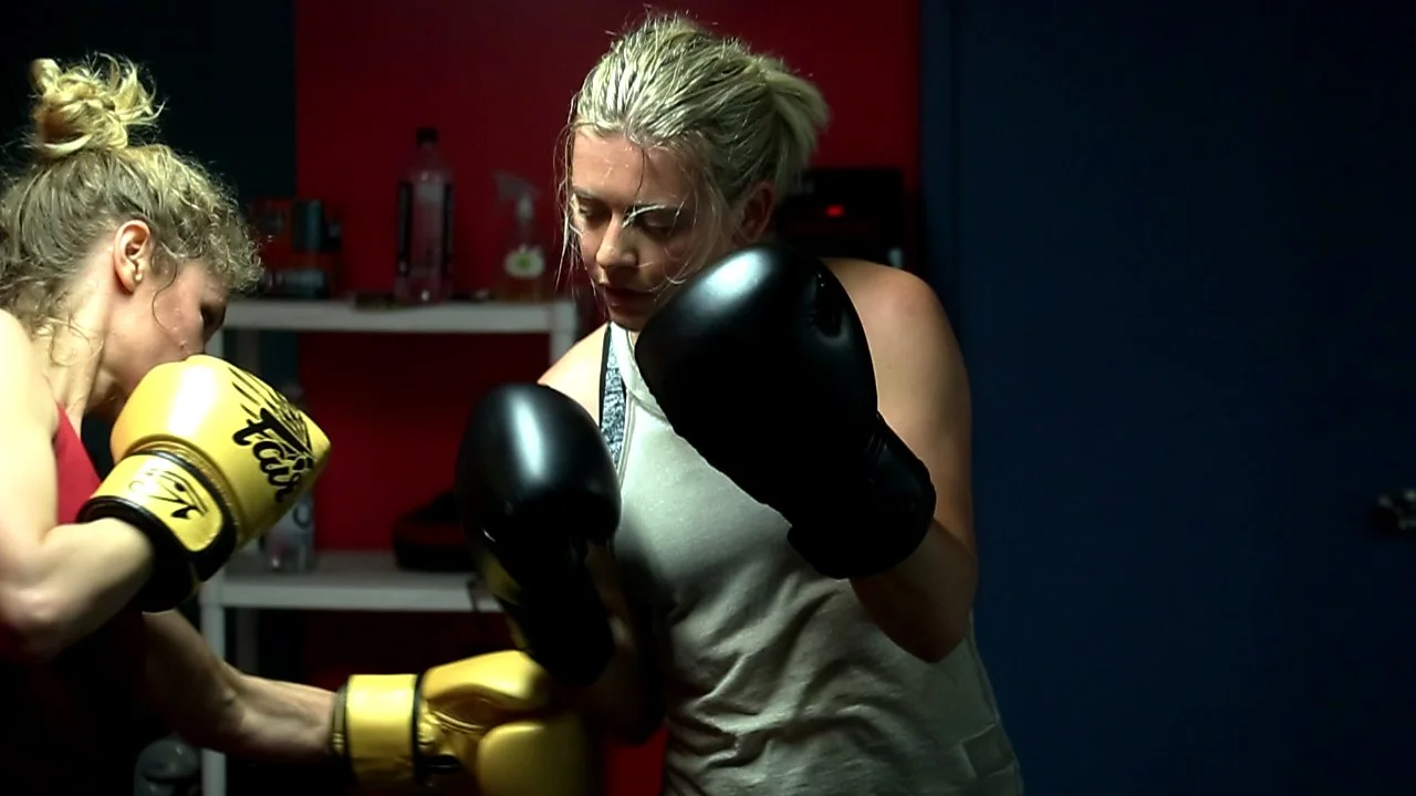 Two women boxing in a gym, wearing boxing gloves, with one woman in yellow gloves and the other in black gloves, in a fighting stance.