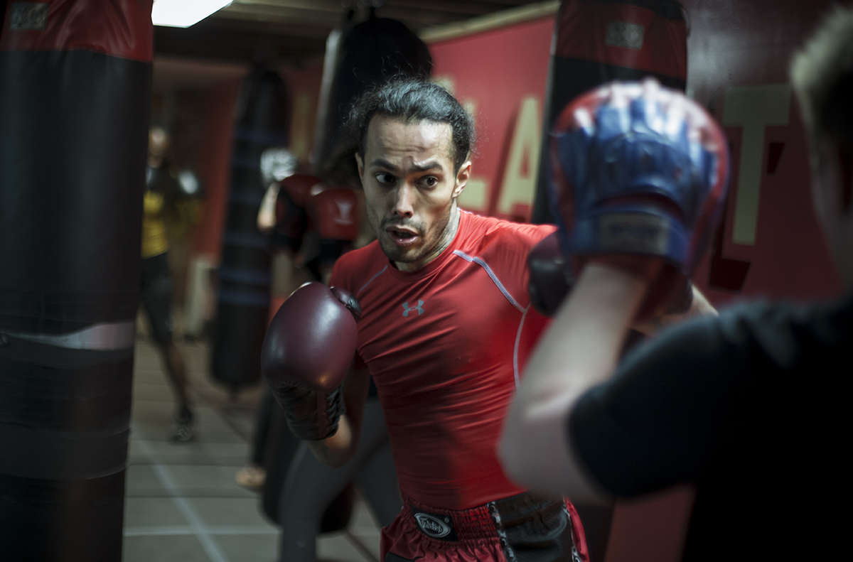 A boxer wearing red workout clothes and boxing gloves practicing punches on a punching bag in a gym, with other gym equipment in the background.