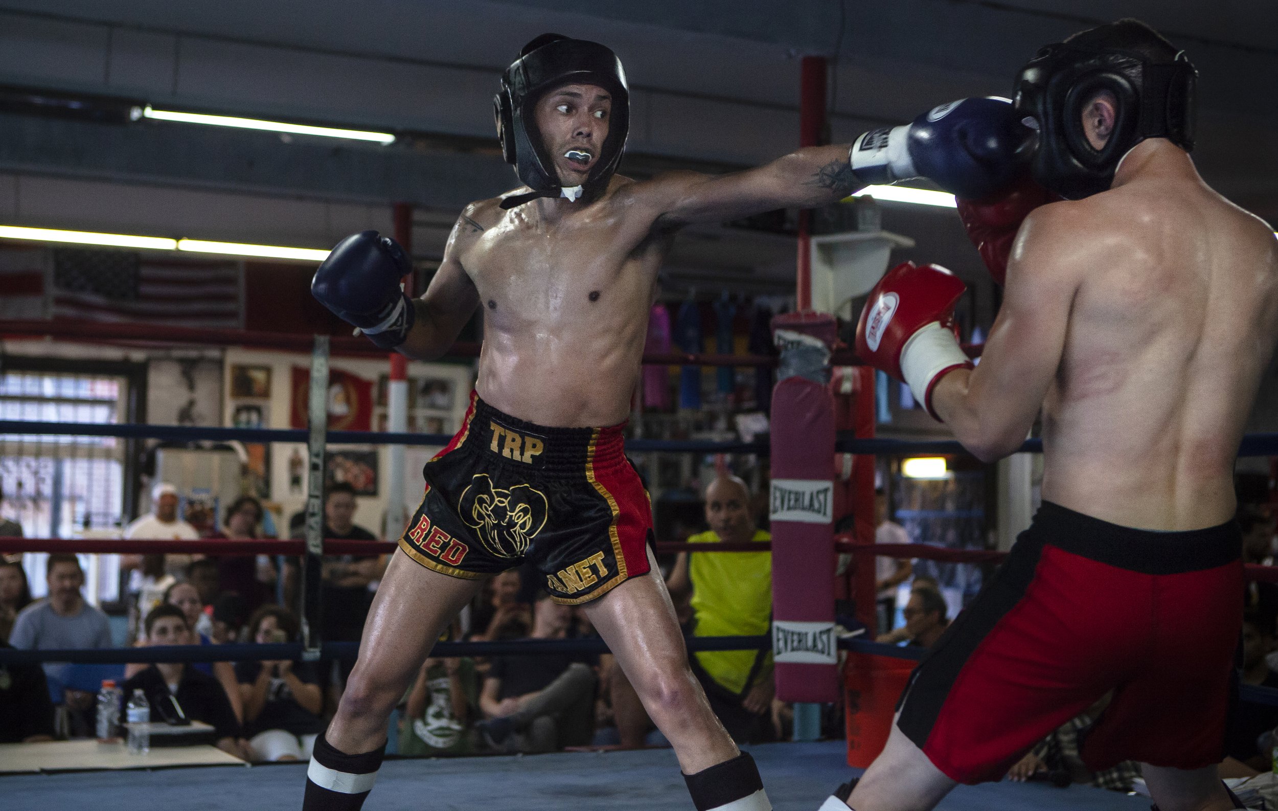 Two shirtless men sparring in a boxing ring with an audience watching in the background.