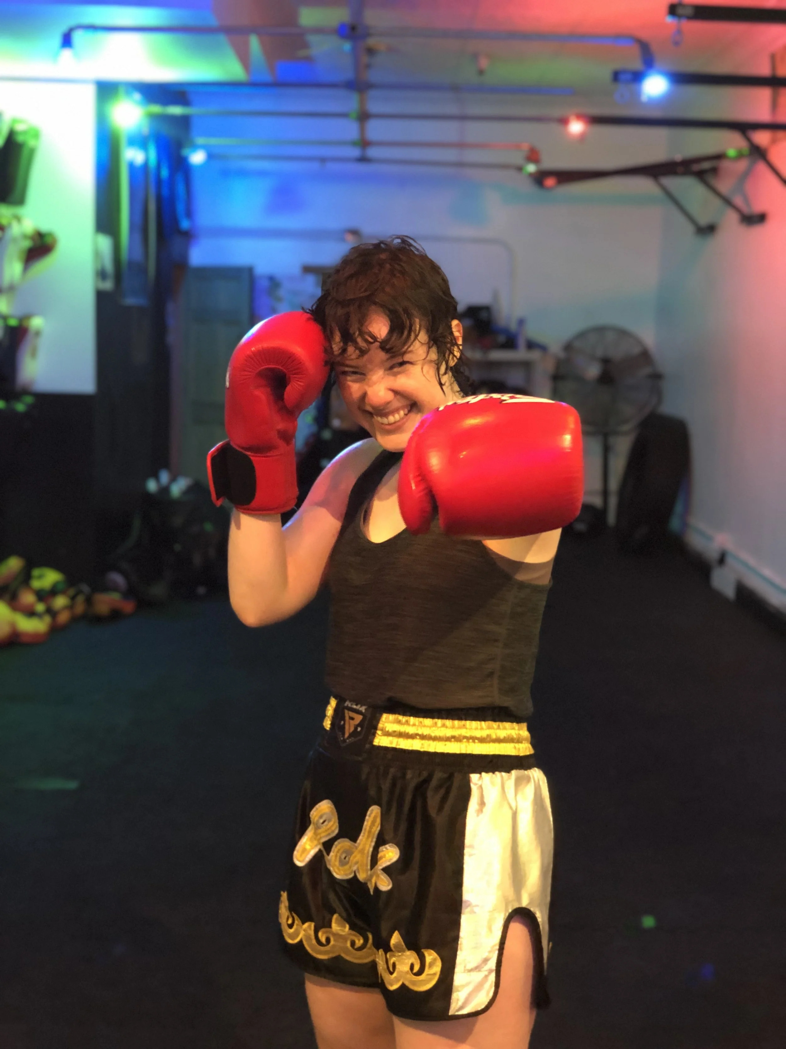 A woman in boxing gear posing with a smile in a boxing gym, wearing red boxing gloves and black and gold shorts.