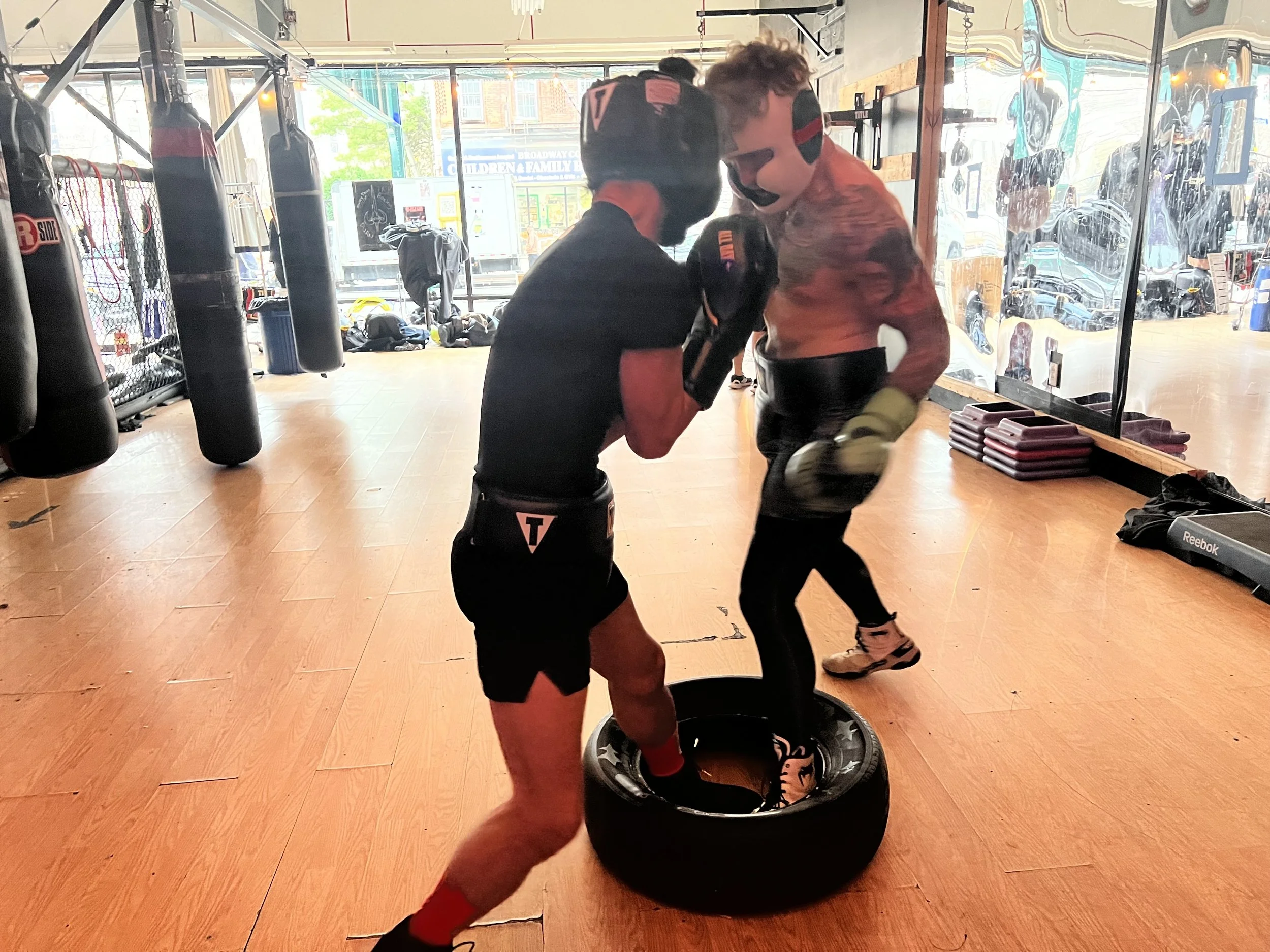 Two people practicing boxing in a gym; one standing inside a tire with boxing gloves, the other striking the gloves from outside the tire.